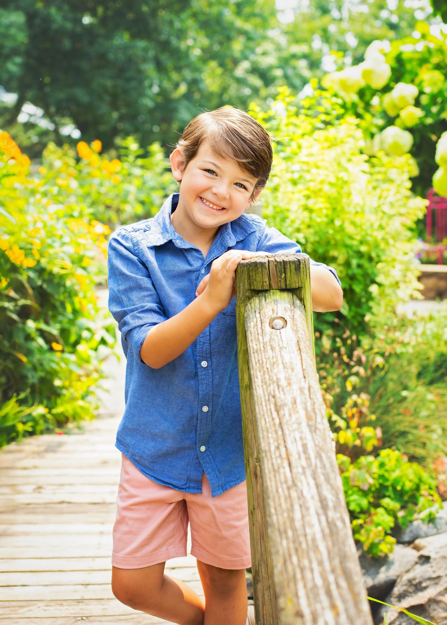 Happy young boy in blue shirt leans on wooden post smiling on sunny garden bridge walkway.