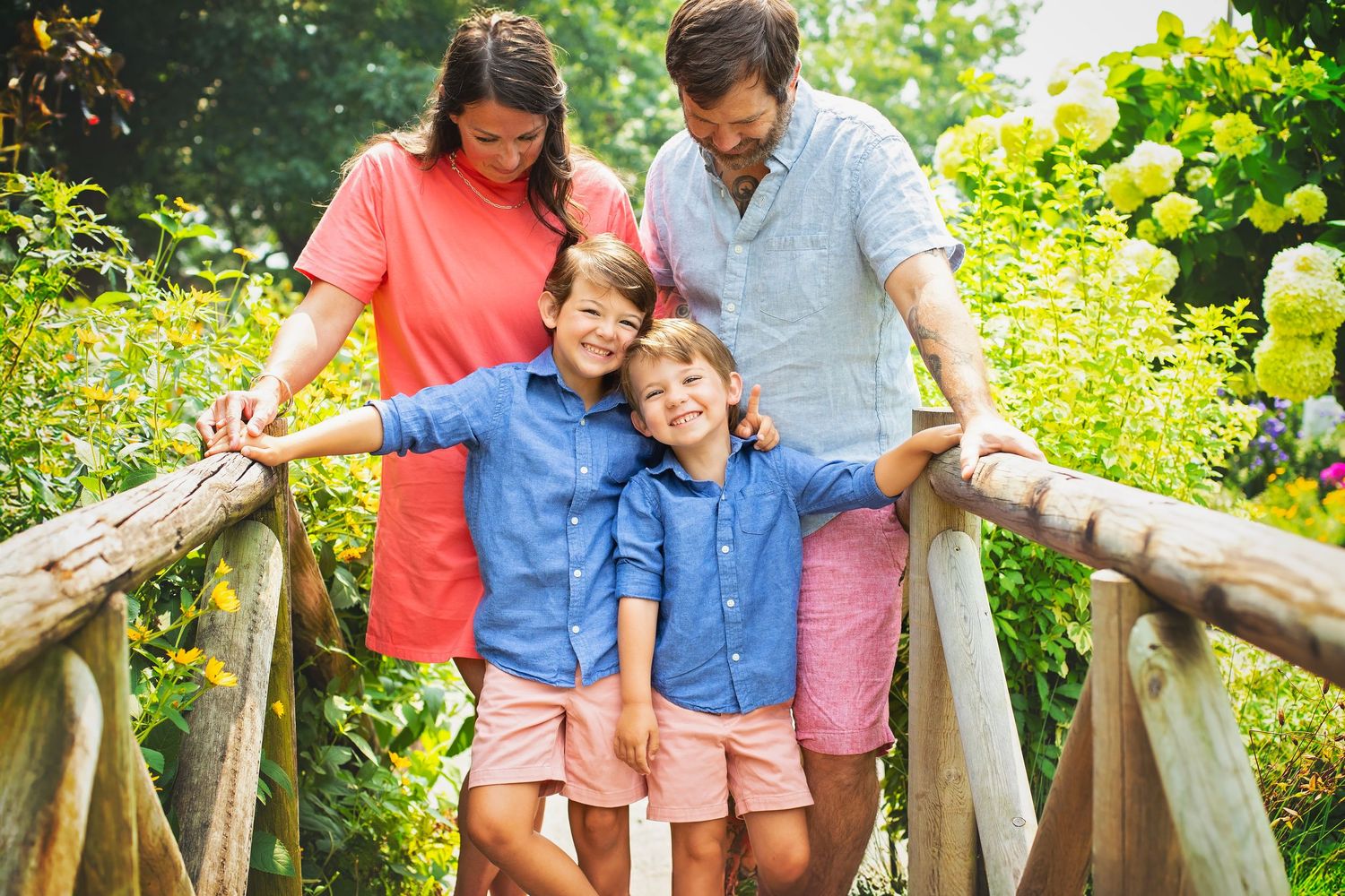 Happy family of four poses on rustic wooden bridge surrounded by lush green garden foliage.