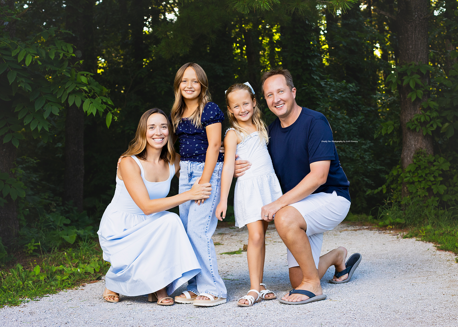 Happy family of four posing outdoors on a gravel path surrounded by lush green trees during a summer photo session.