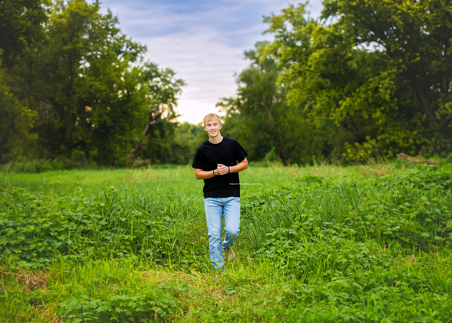 Young man in black shirt and jeans walking through lush green field surrounded by trees on a cloudy day.