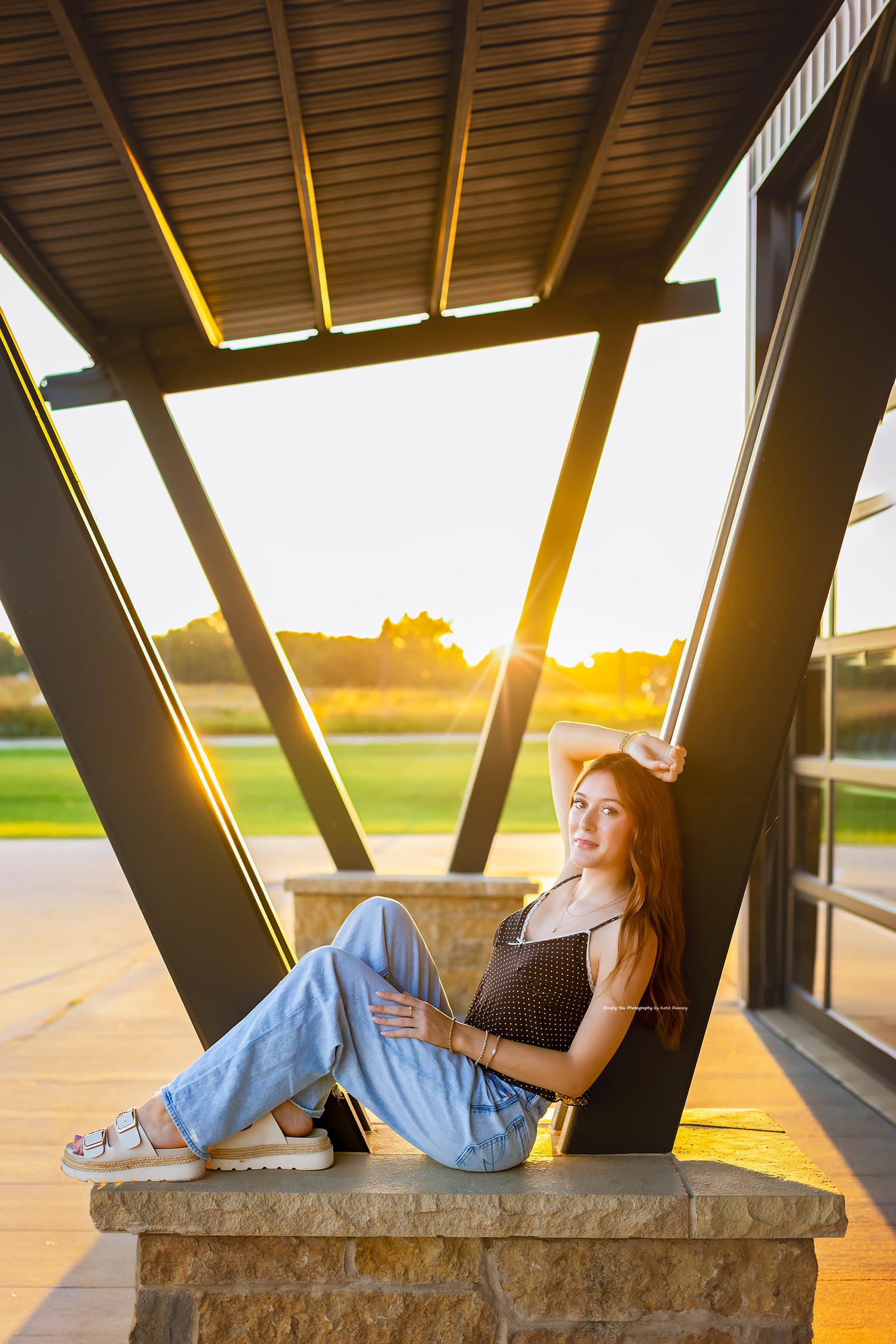 Young woman in jeans and black top poses at golden hour under modern architectural beams with sunlight streaming through.