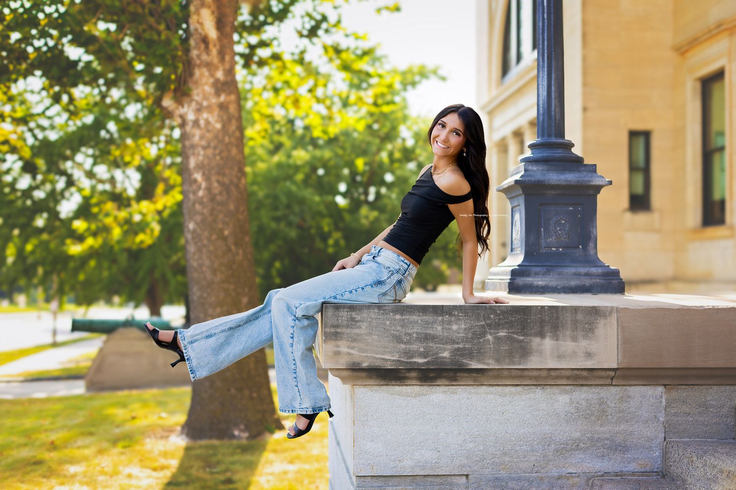 Young woman in black top and wide-leg jeans posing on a stone ledge near a lamppost in a sunny park.