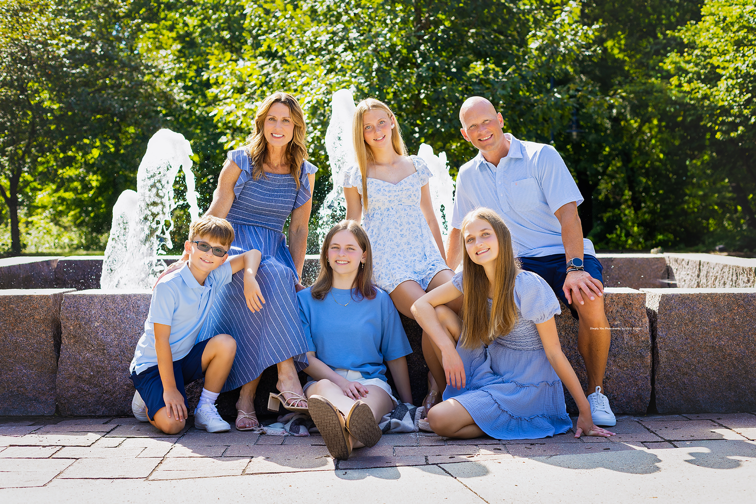 Happy family of six posing together outdoors near a fountain on a sunny day, wearing coordinated blue outfits.