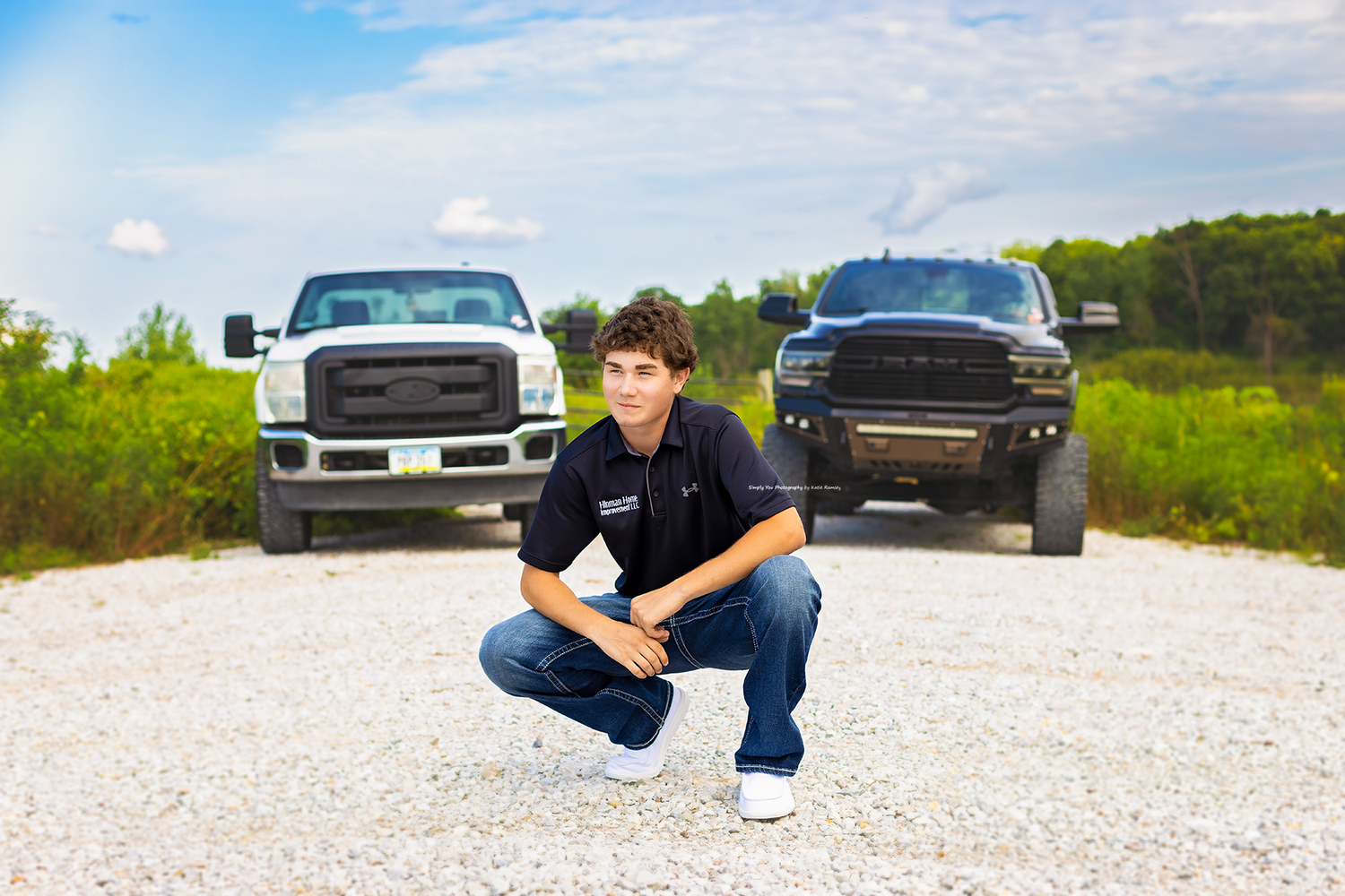 Teen boy crouching between two trucks on gravel road, white Ford F-250 and black Ram pickup in background.