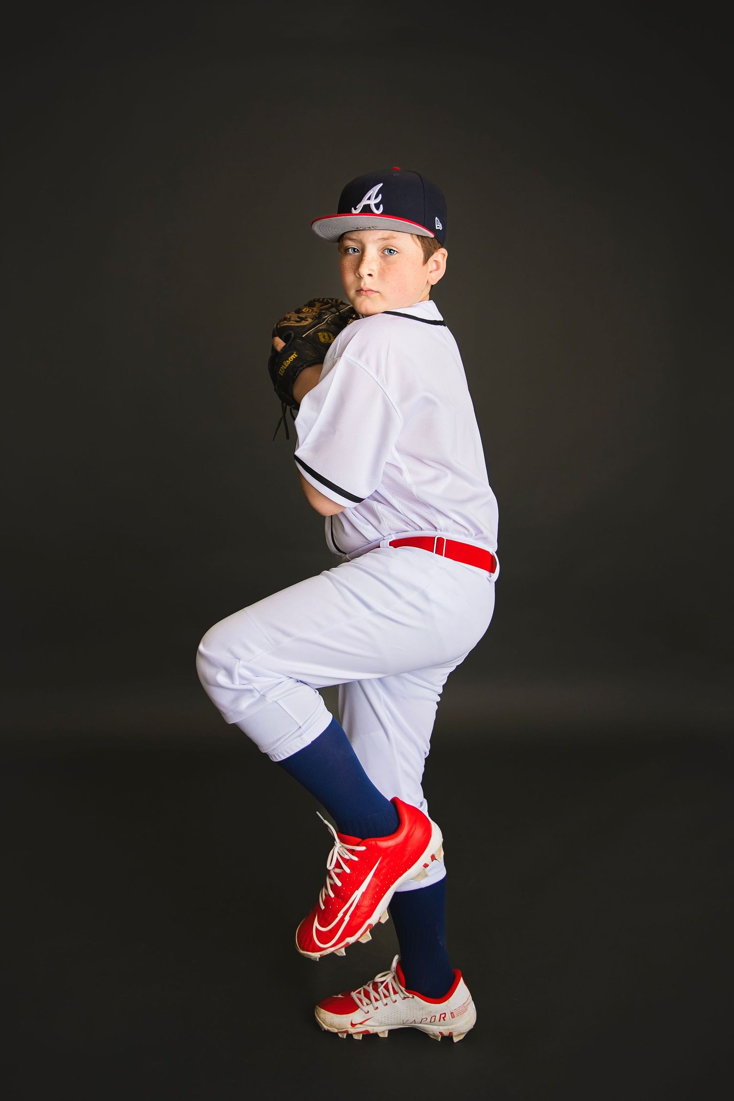 Youth baseball pitcher in Braves uniform mid-windup stance, wearing red cleats, dark studio background.