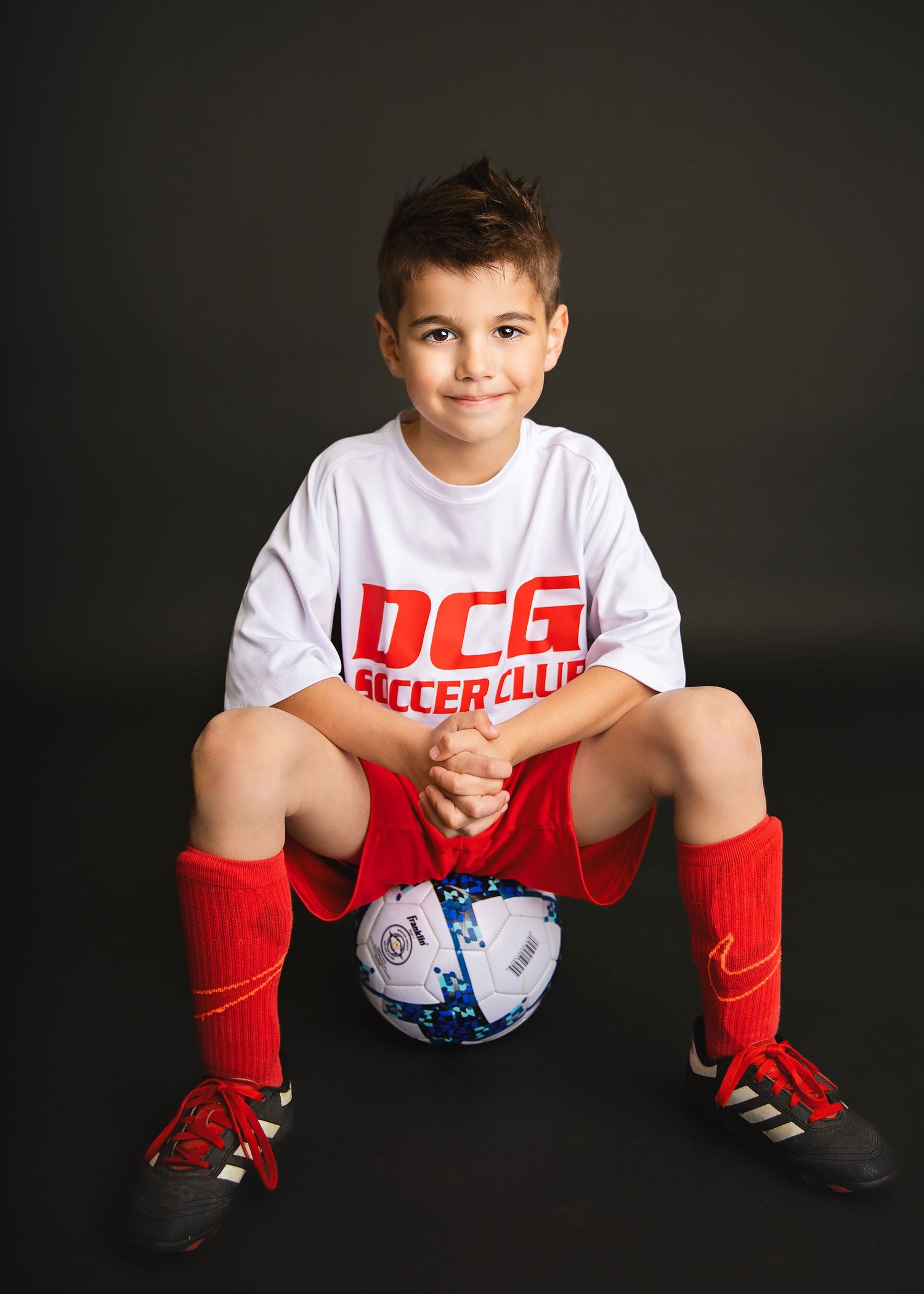 Young boy in DCG Soccer Club white jersey and red shorts sits on soccer ball smiling at camera.
