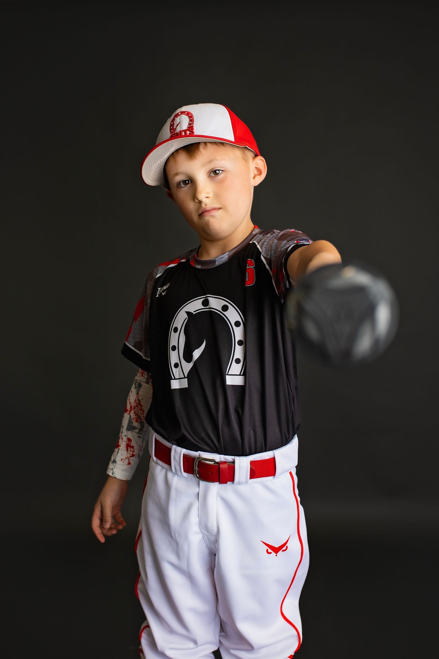 Young baseball player in black horseshoe jersey extends glove toward camera in studio portrait.