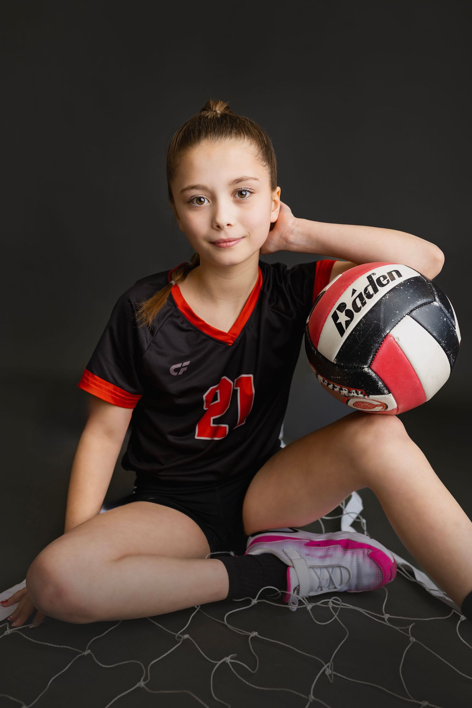 Young girl in number 21 volleyball uniform sitting cross-legged holding Baden volleyball against dark background.