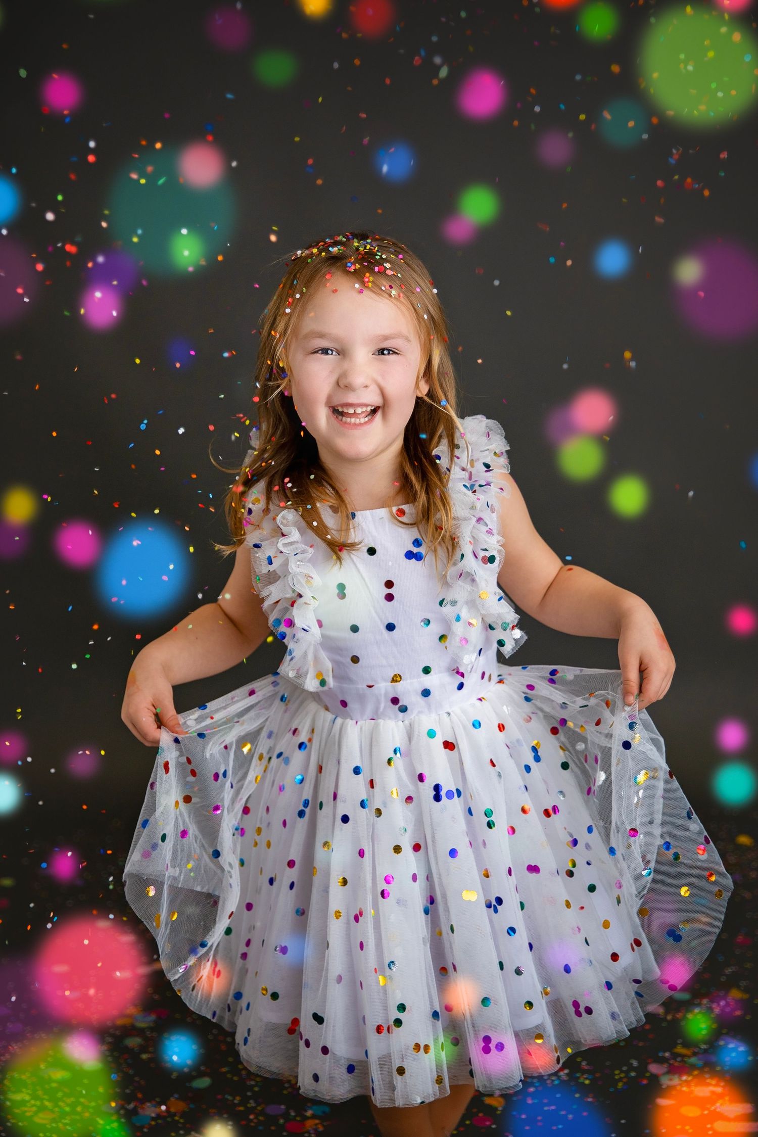 A happy young girl in a white polka dot dress twirls and laughs surrounded by colorful bokeh and confetti.