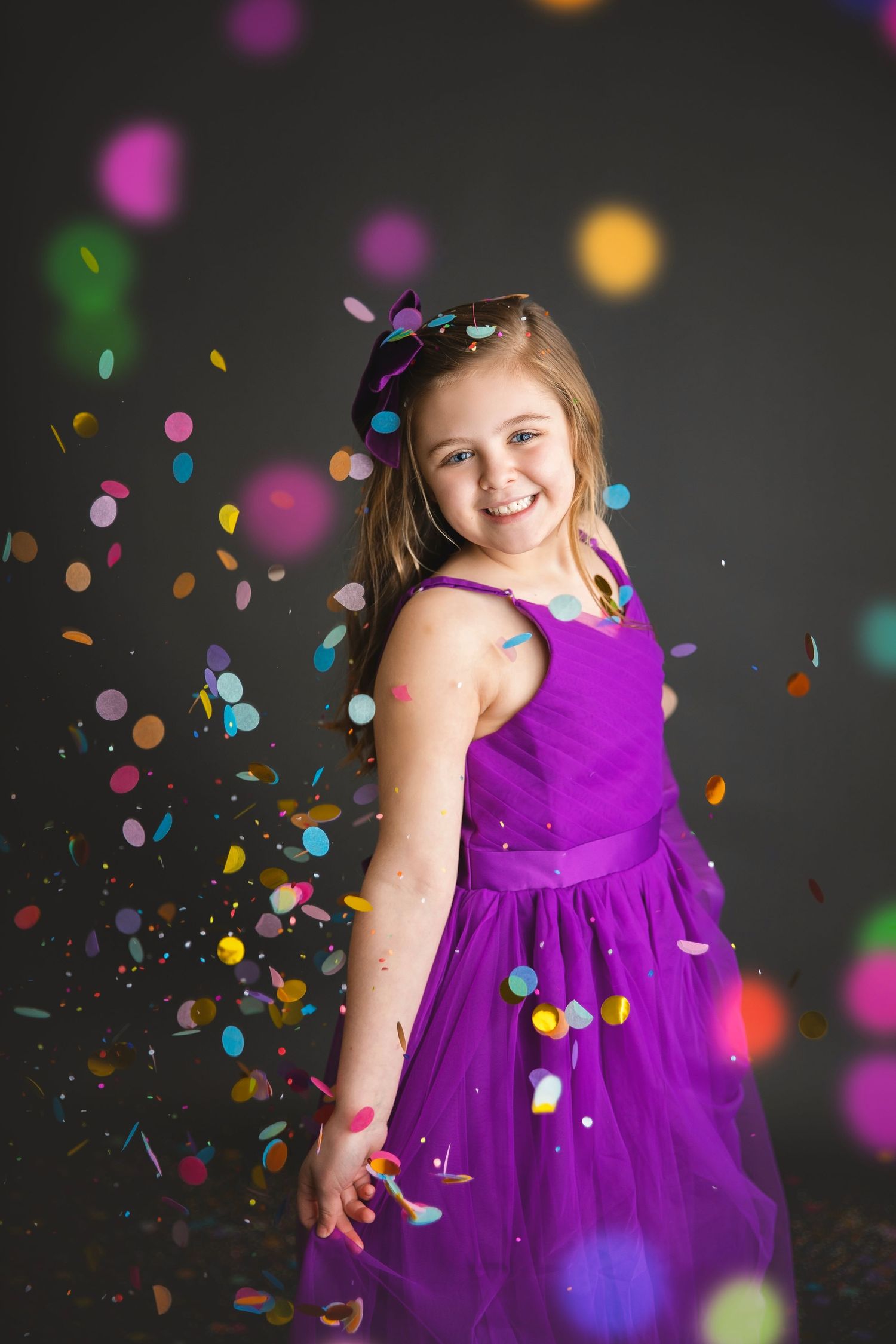 Happy girl in purple tulle dress twirling amid falling colorful confetti in a studio photo shoot.
