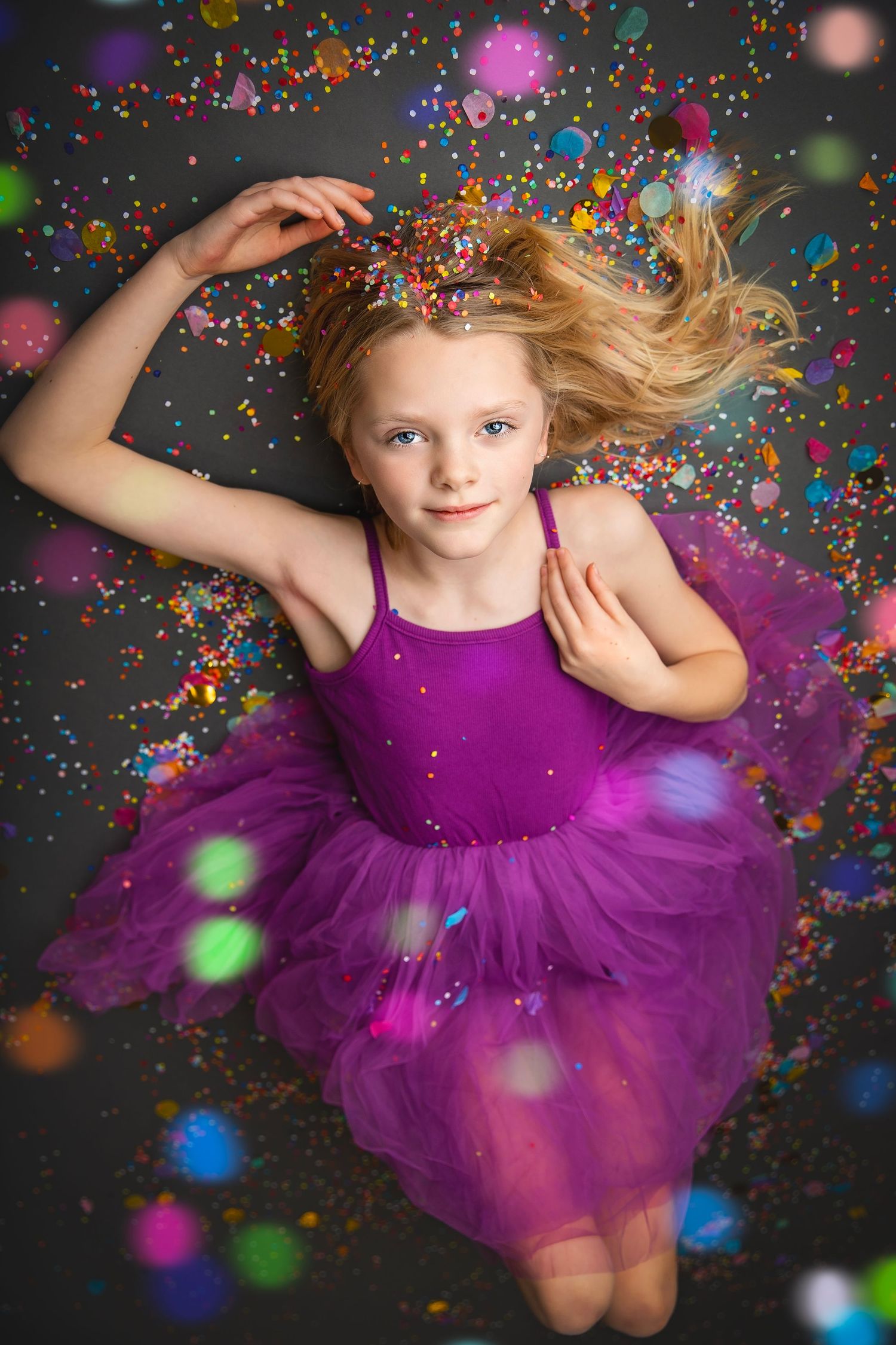 Blonde girl in purple tutu lying down surrounded by colorful bokeh confetti in studio portrait.
