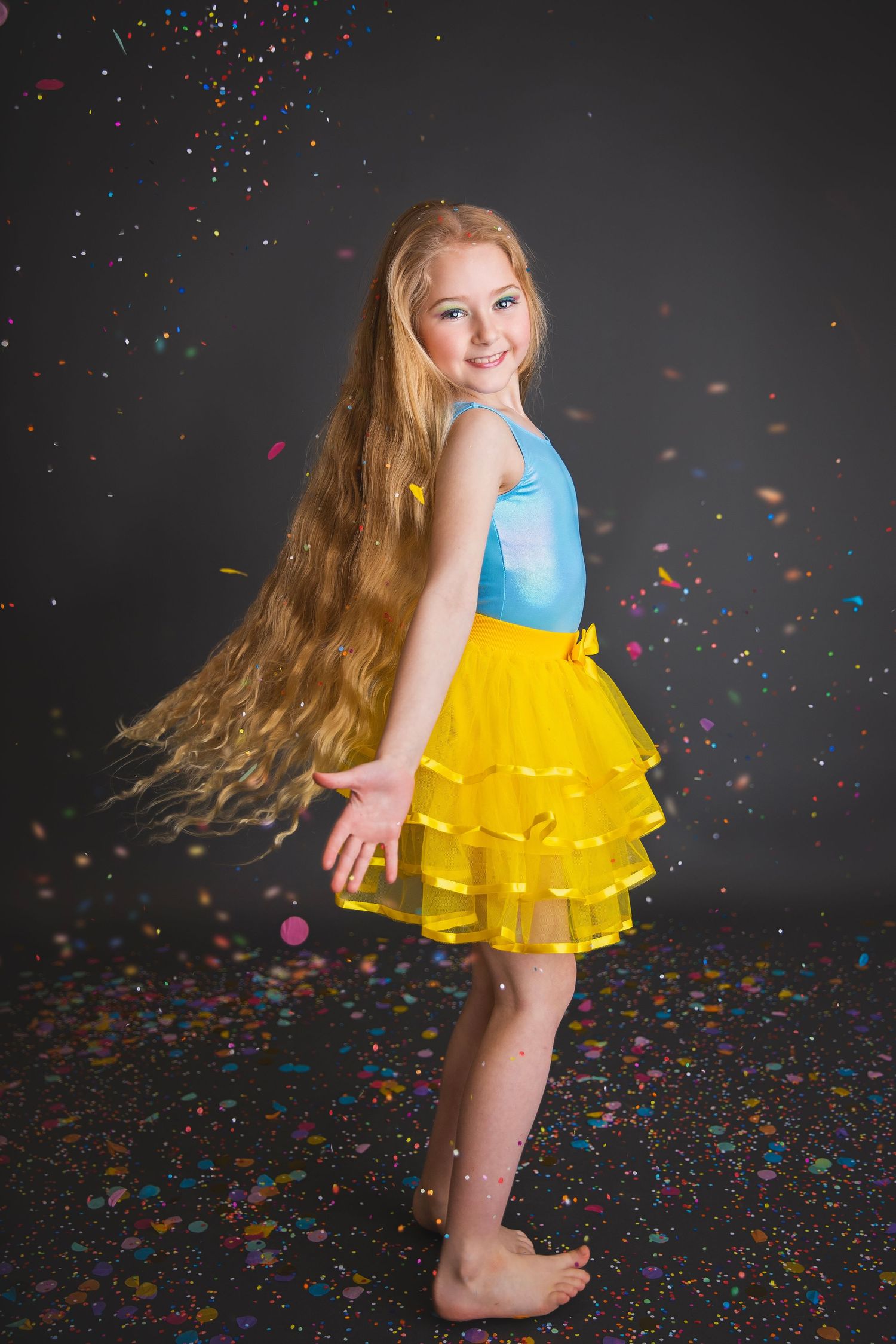 A young girl with long blonde hair twirls in a blue top and yellow tutu skirt with colorful confetti falling around her.