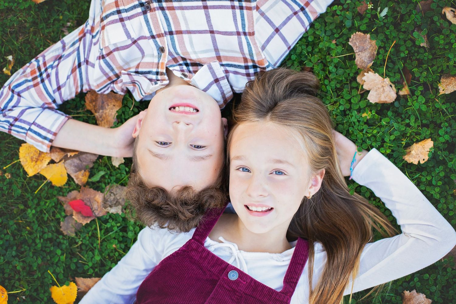 Two children lying on grass surrounded by colorful autumn leaves smiling.