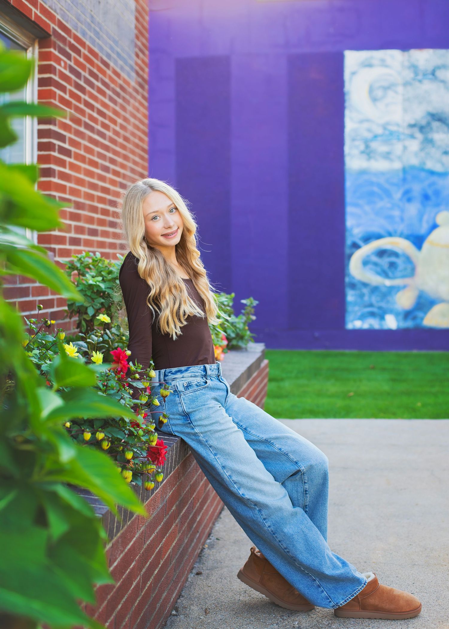Blonde teen in brown top and wide-leg jeans sitting by flowers near purple wall.