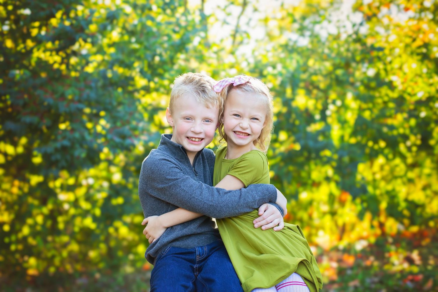 Two young blonde siblings hugging and smiling outdoors with bright green fall foliage.