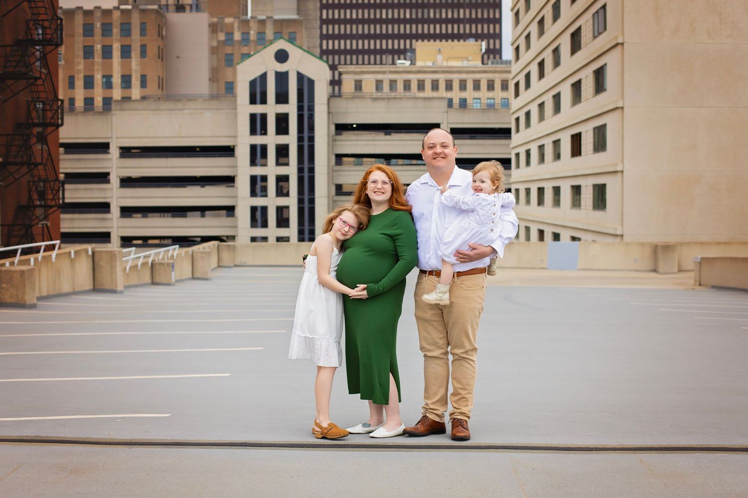 Family of four with pregnant mother in green dress posing on urban rooftop parking garage.