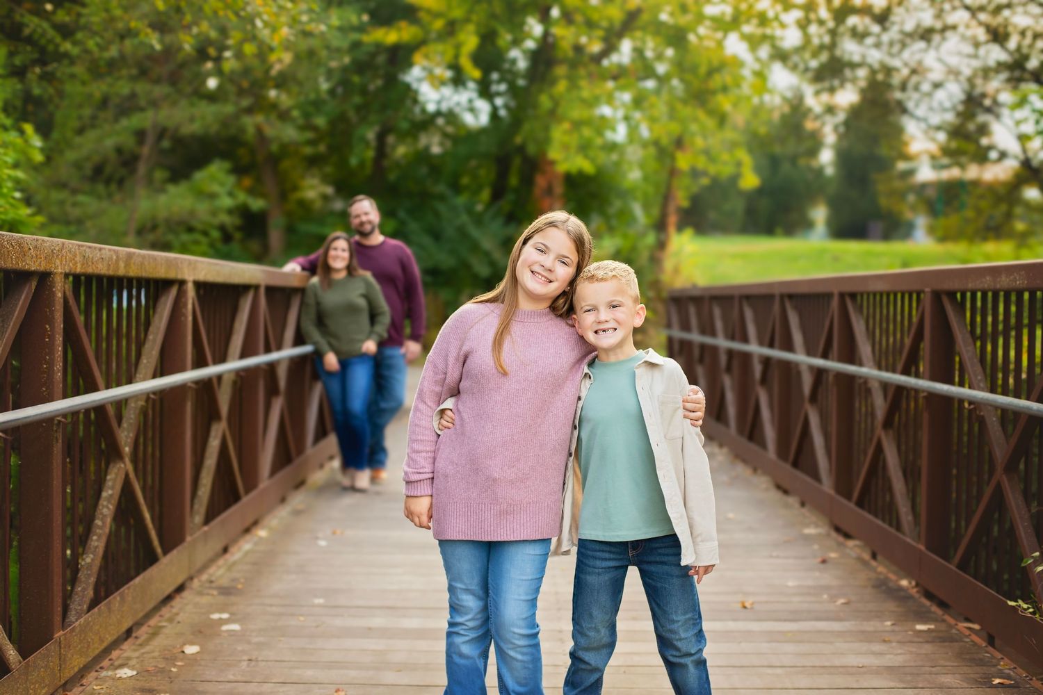 Two siblings hug on a wooden bridge while their parents walk behind them.