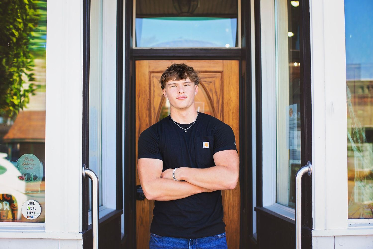Young man in black Carhartt shirt stands with arms crossed in front of a wooden door entrance.