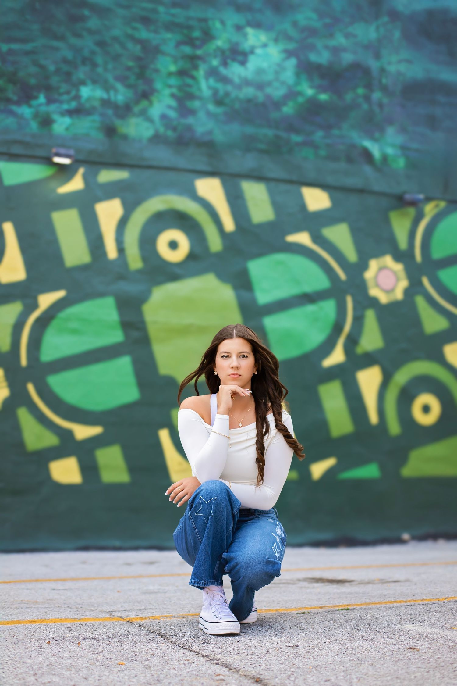 Teen girl in white off-shoulder top and jeans crouches before vibrant green geometric graffiti mural.