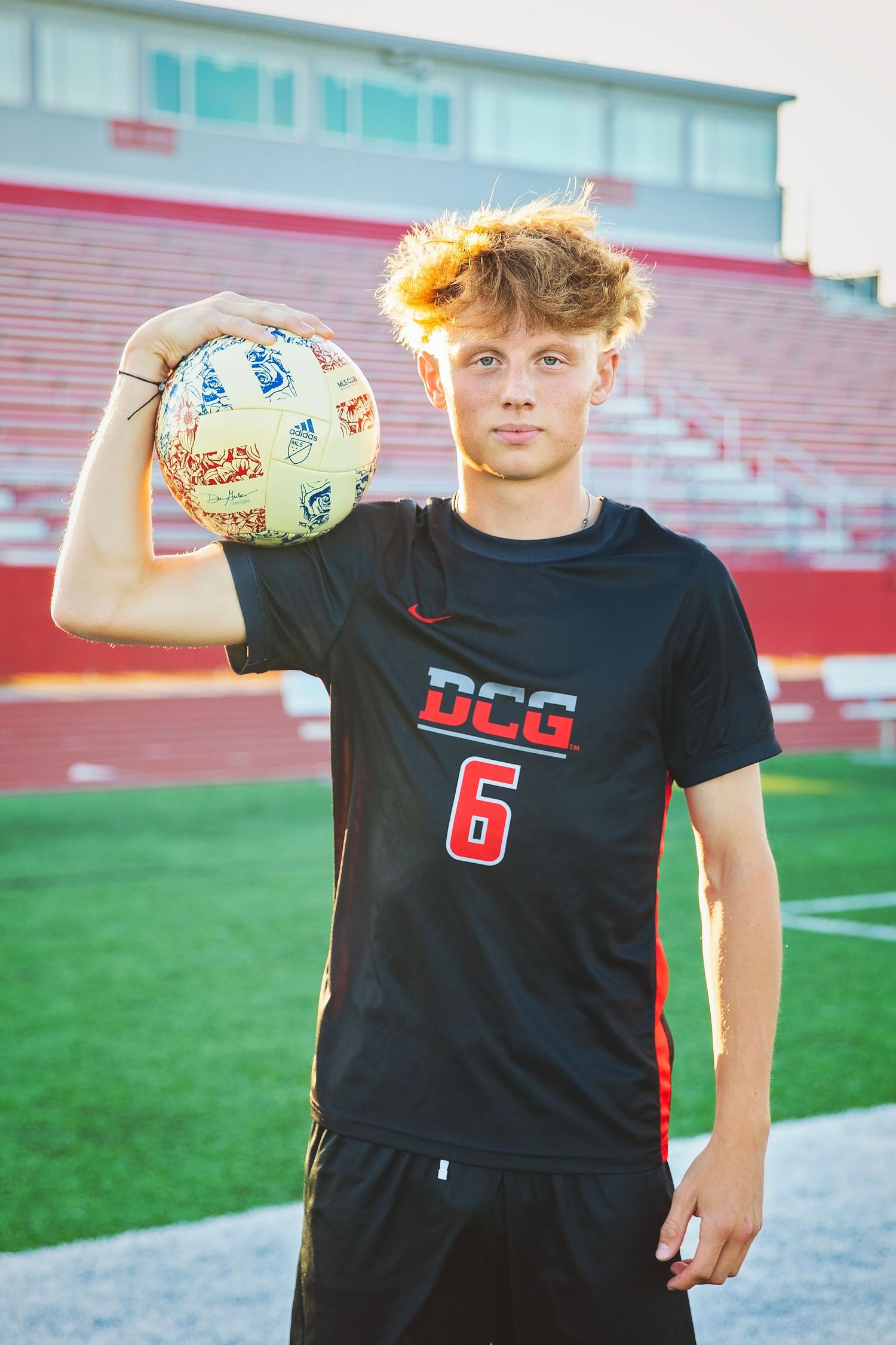 Teen athlete in DCG number 6 soccer jersey holding ball at stadium during golden hour.