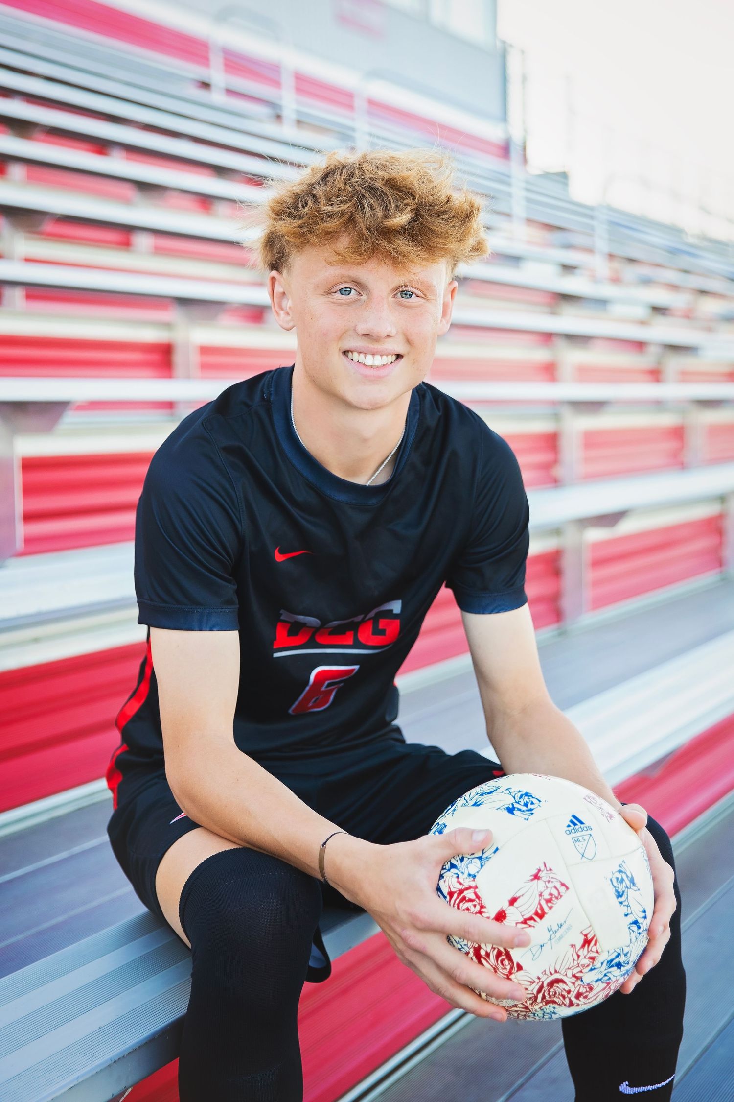 Smiling teen boy holding soccer ball sitting in red stadium bleachers wearing black Nike jersey.
