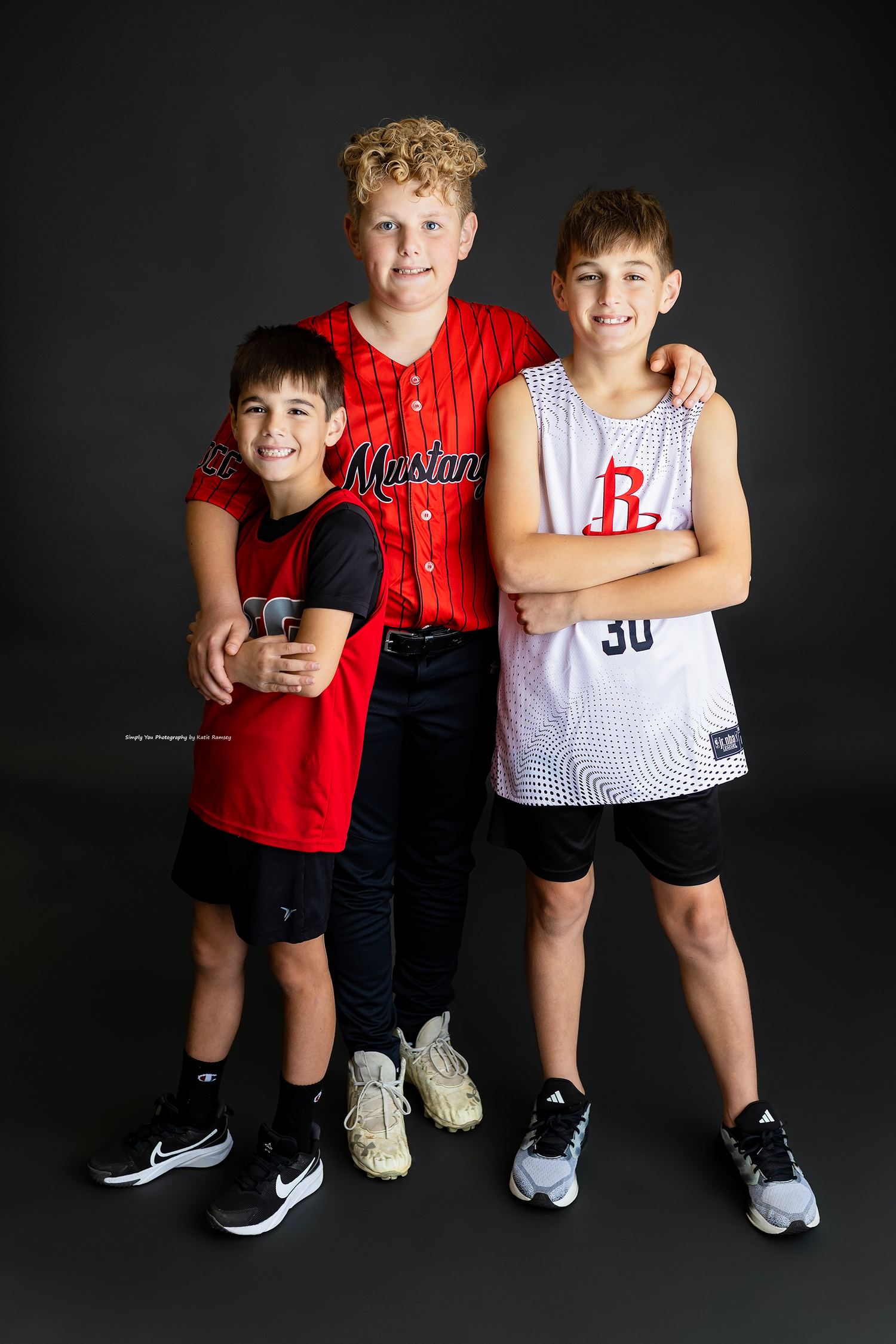 Three boys in sports jerseys pose together smiling in a studio portrait setting.