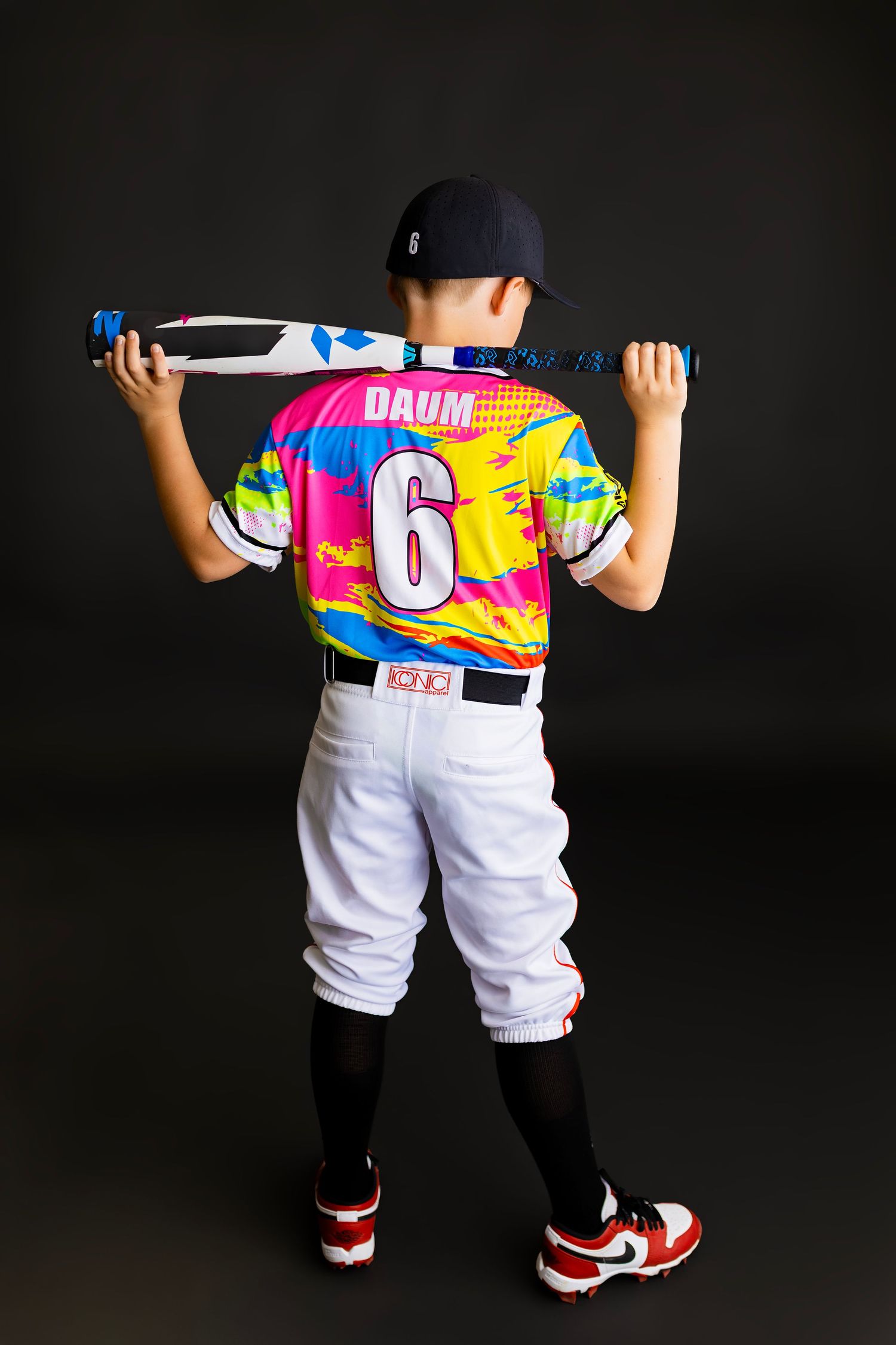 Young baseball player wearing colorful jersey number 6 with DAUM name, white pants, holding bat across shoulders.