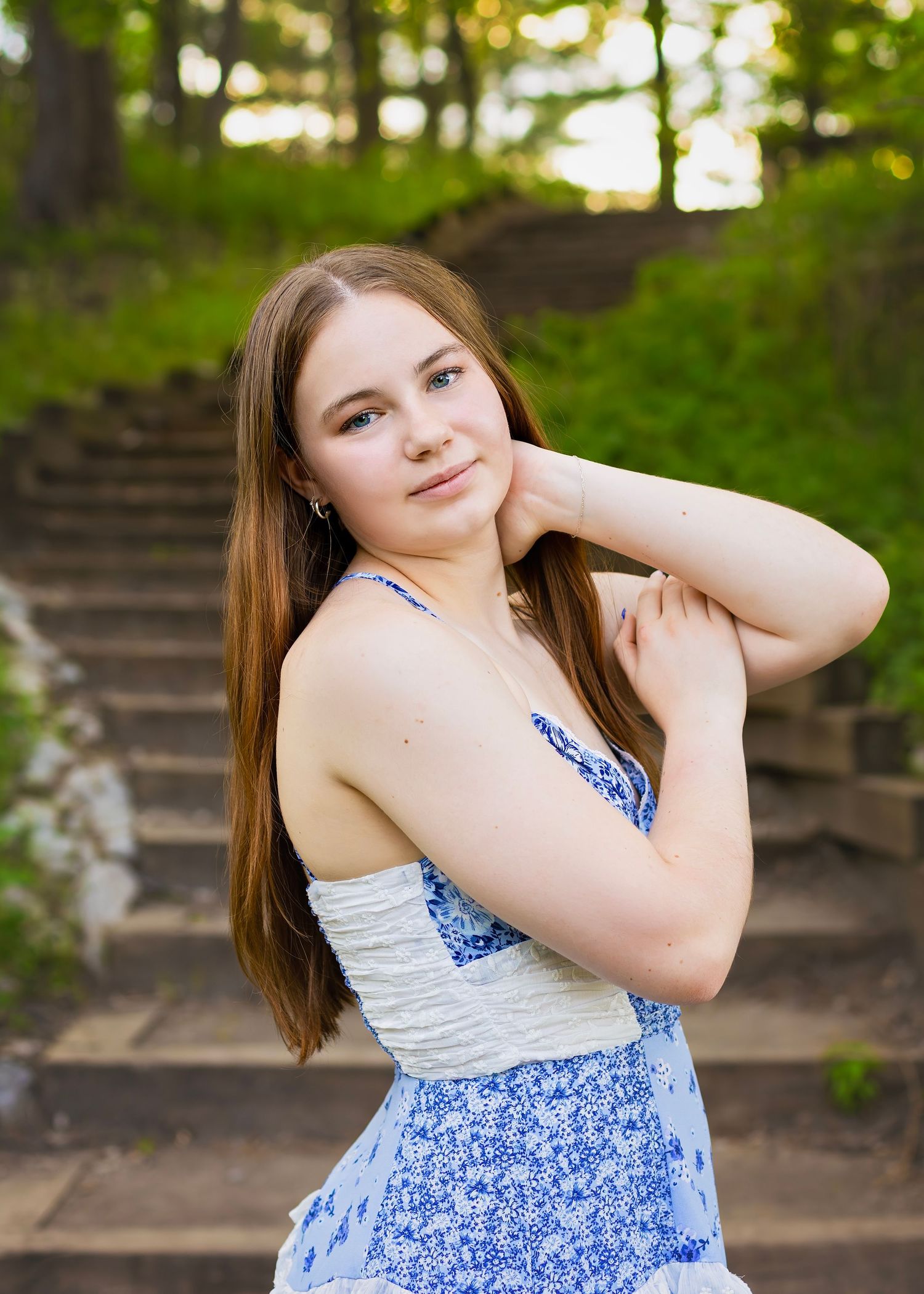 Teenage girl in blue floral sundress posing on stone steps in wooded park setting.
