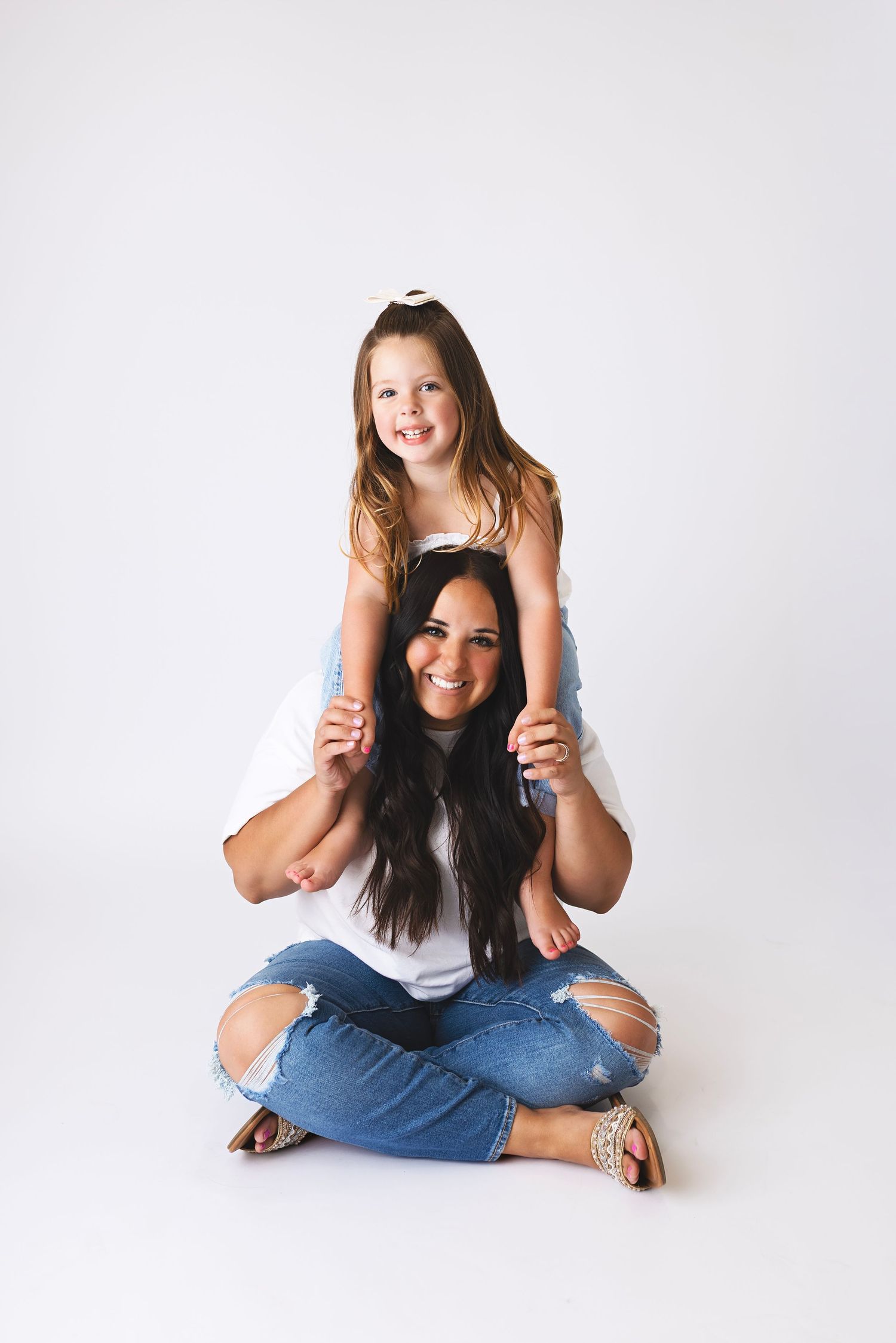 A woman with long dark hair sits cross-legged on white background with a young girl sitting on her shoulders, both smiling.