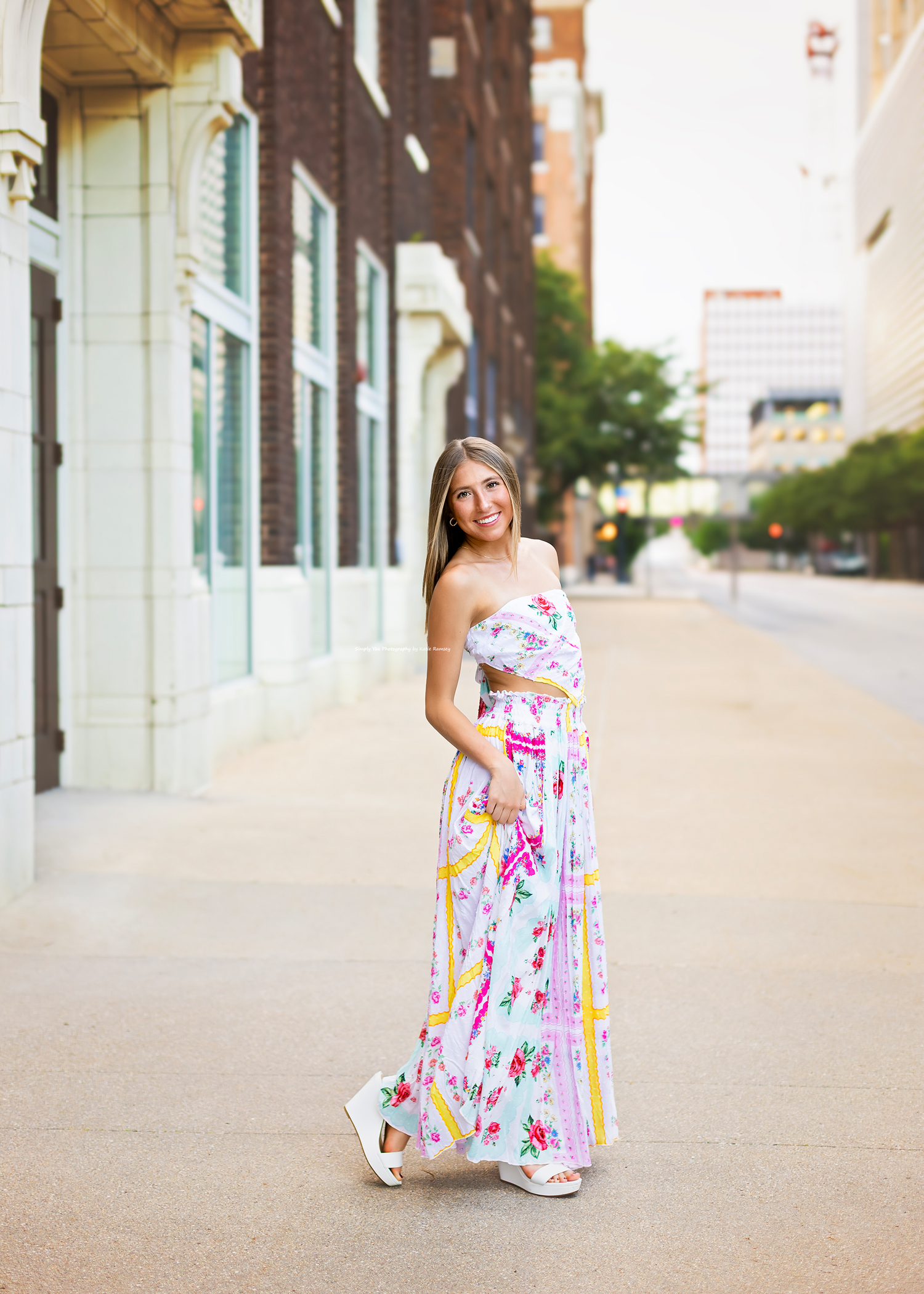 Young woman in colorful floral maxi dress walking on urban sidewalk in downtown setting.