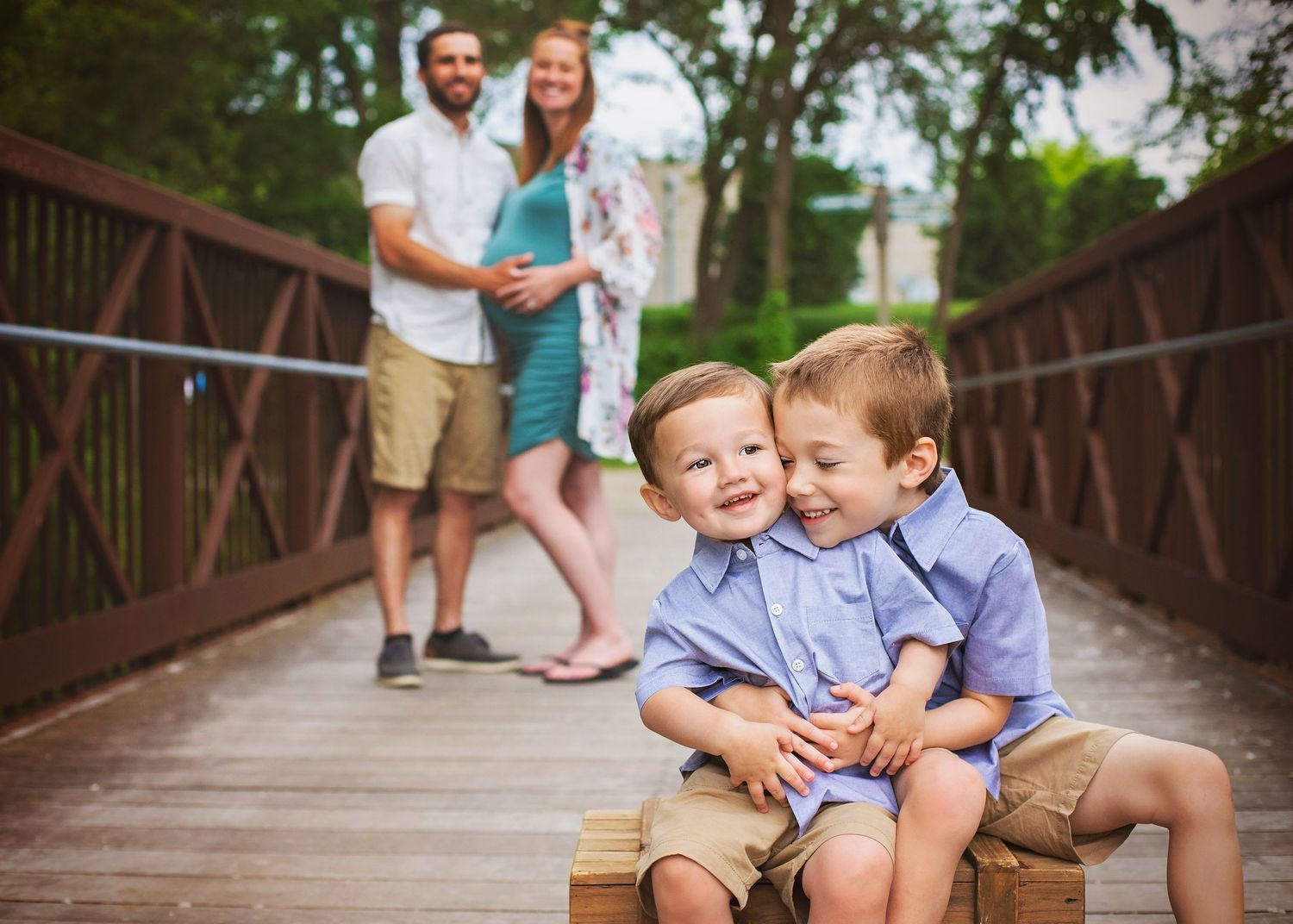Two young brothers smiling on a bridge with expecting parents in the background.