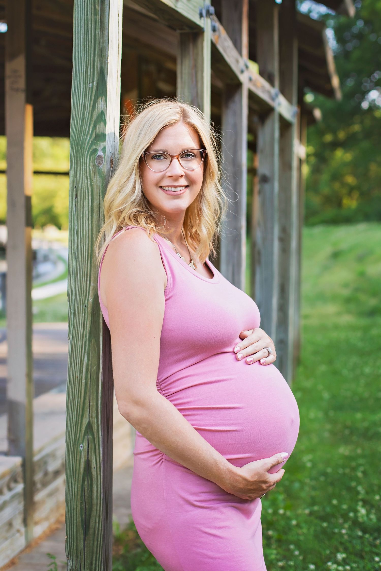 Smiling pregnant woman in pink dress and glasses leaning against wooden post outdoors.