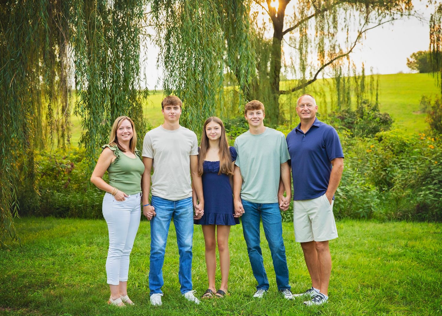 A happy family of five poses outdoors on green grass beneath a weeping willow tree in golden hour light.