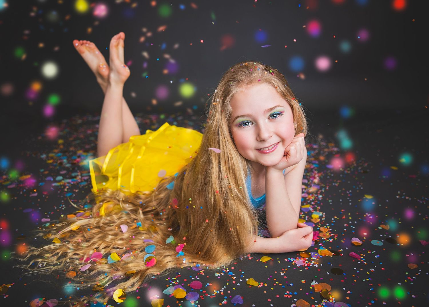 Smiling girl in yellow tutu lying on floor surrounded by colorful confetti in studio setting.