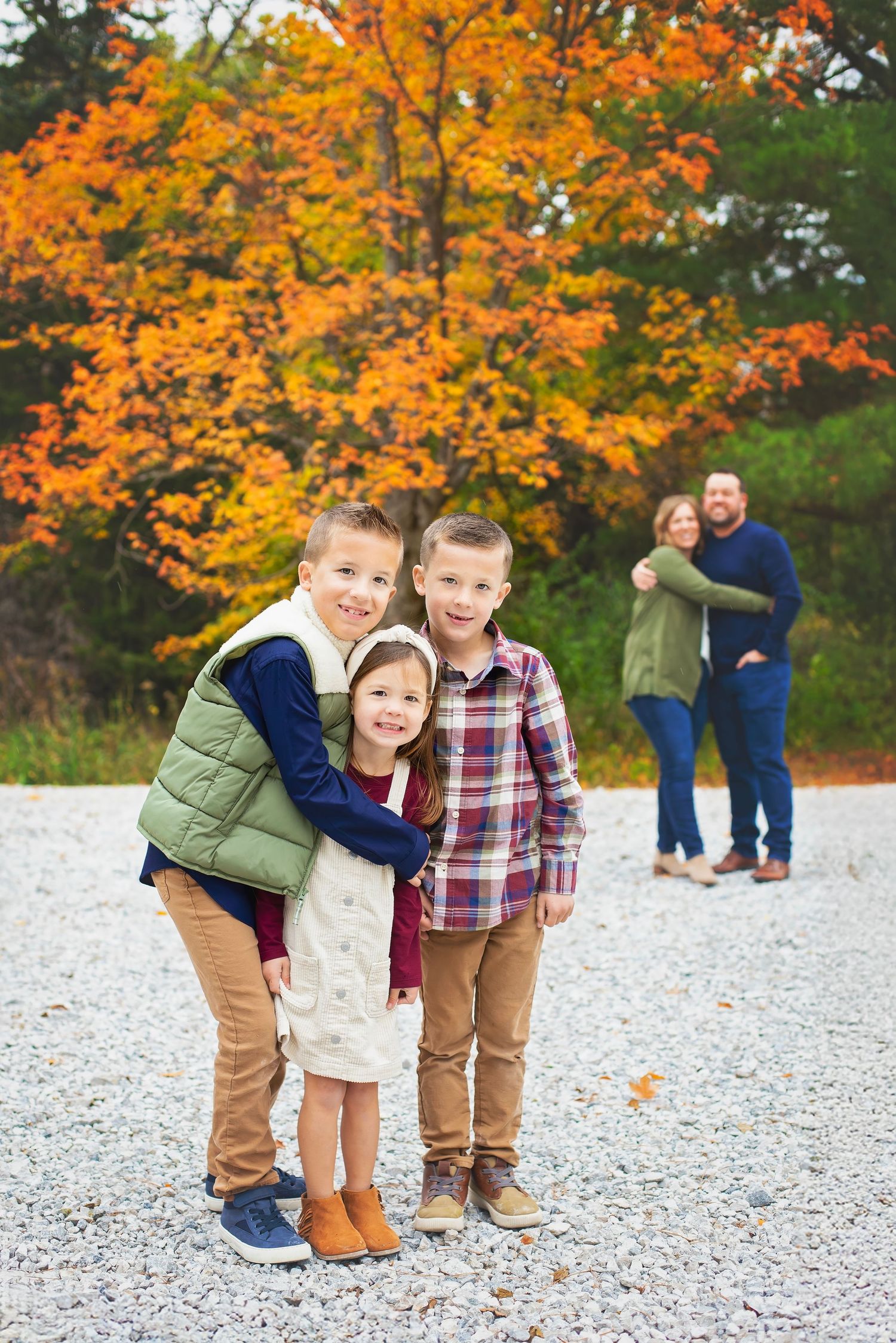 Three siblings hug and smile outdoors with vibrant orange fall foliage while parents embrace in the background.