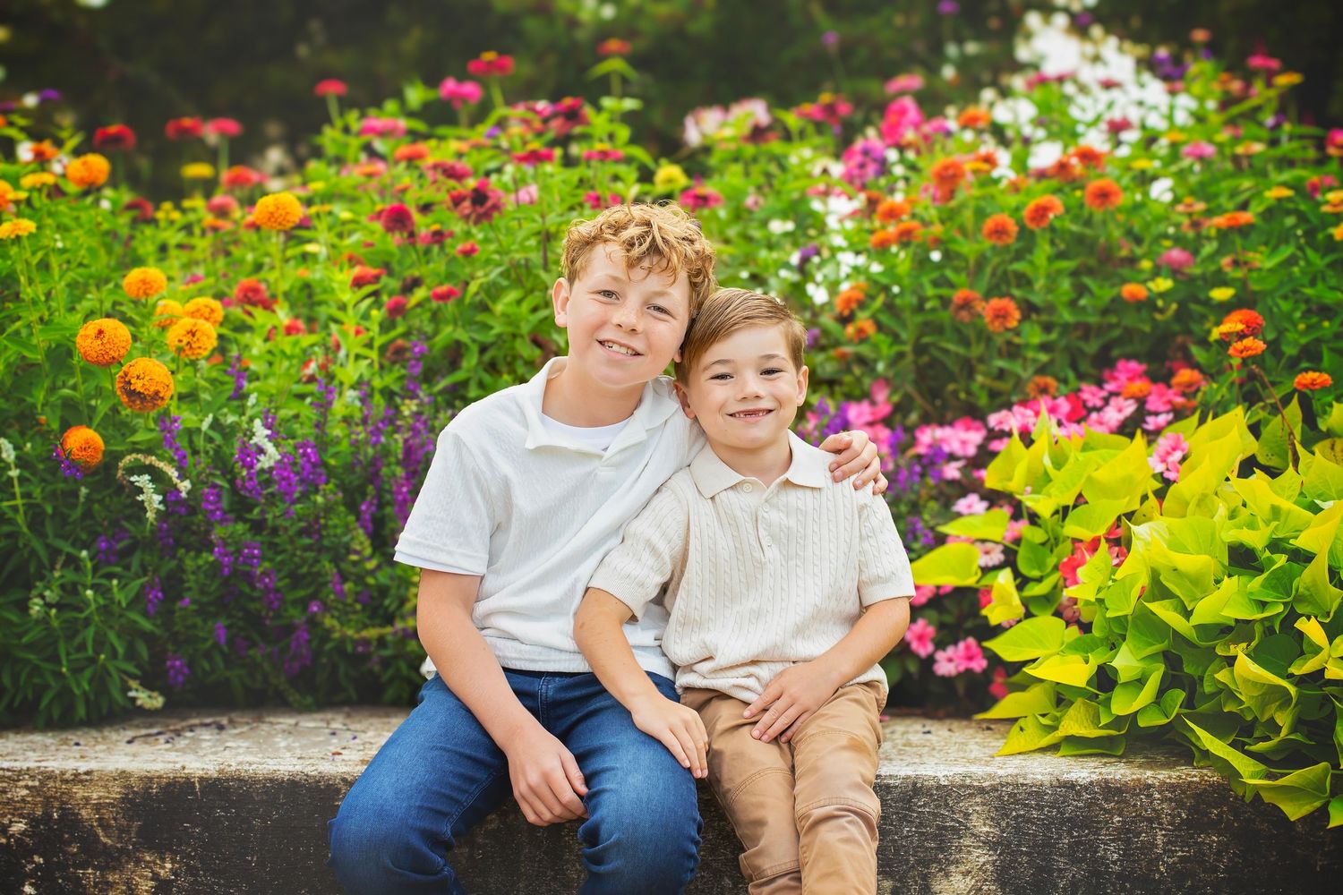 Two smiling brothers sitting together in front of a vibrant colorful flower garden outdoors.