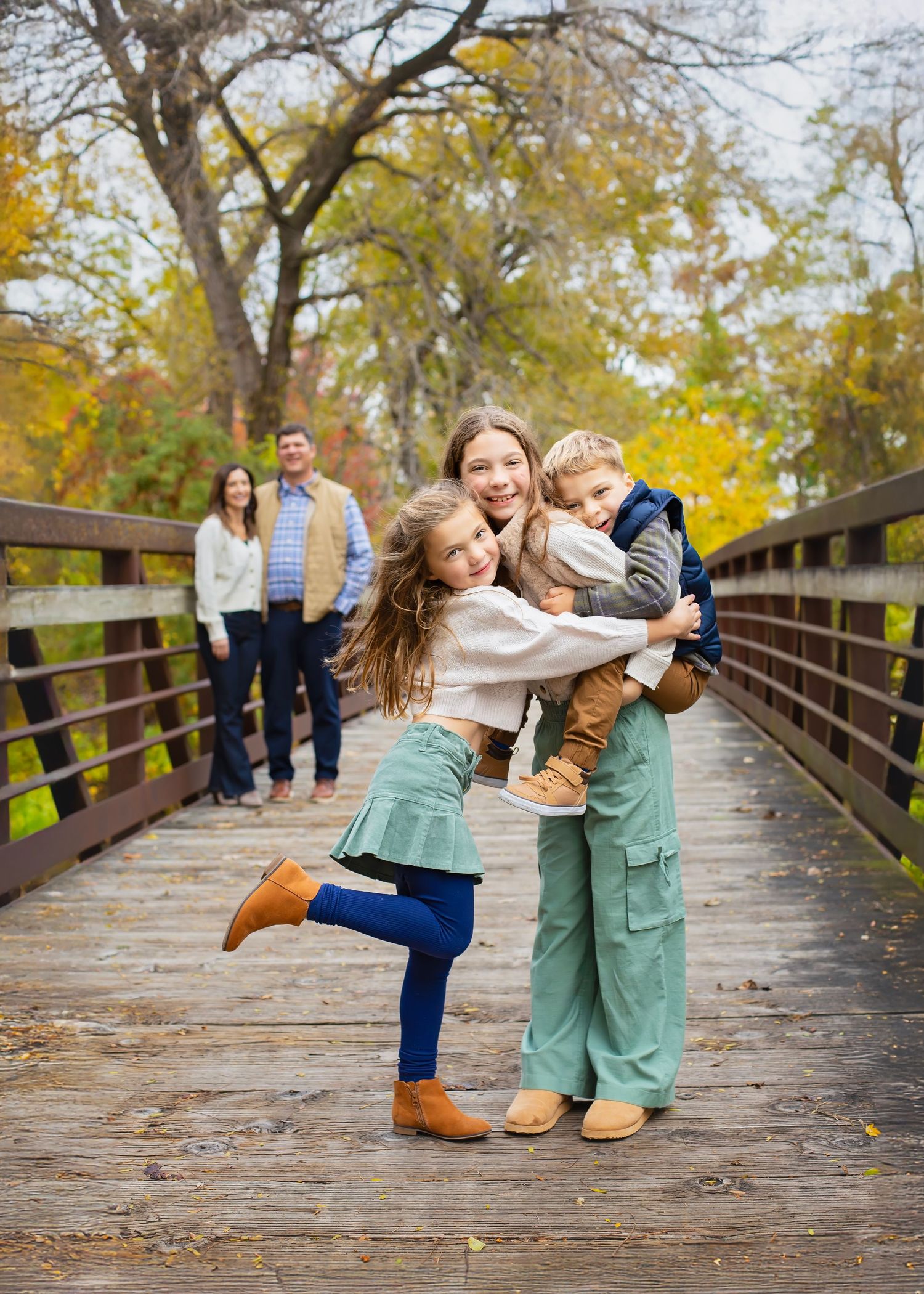 Teenage girl hugging younger siblings on a wooden bridge with parents in background.