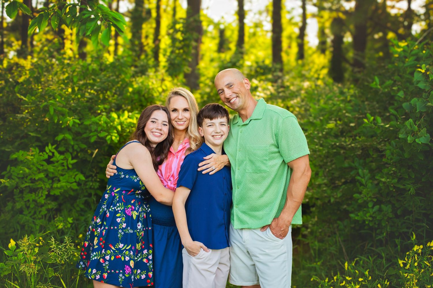 Family of four hugging and smiling together outdoors surrounded by green trees in warm sunlight.
