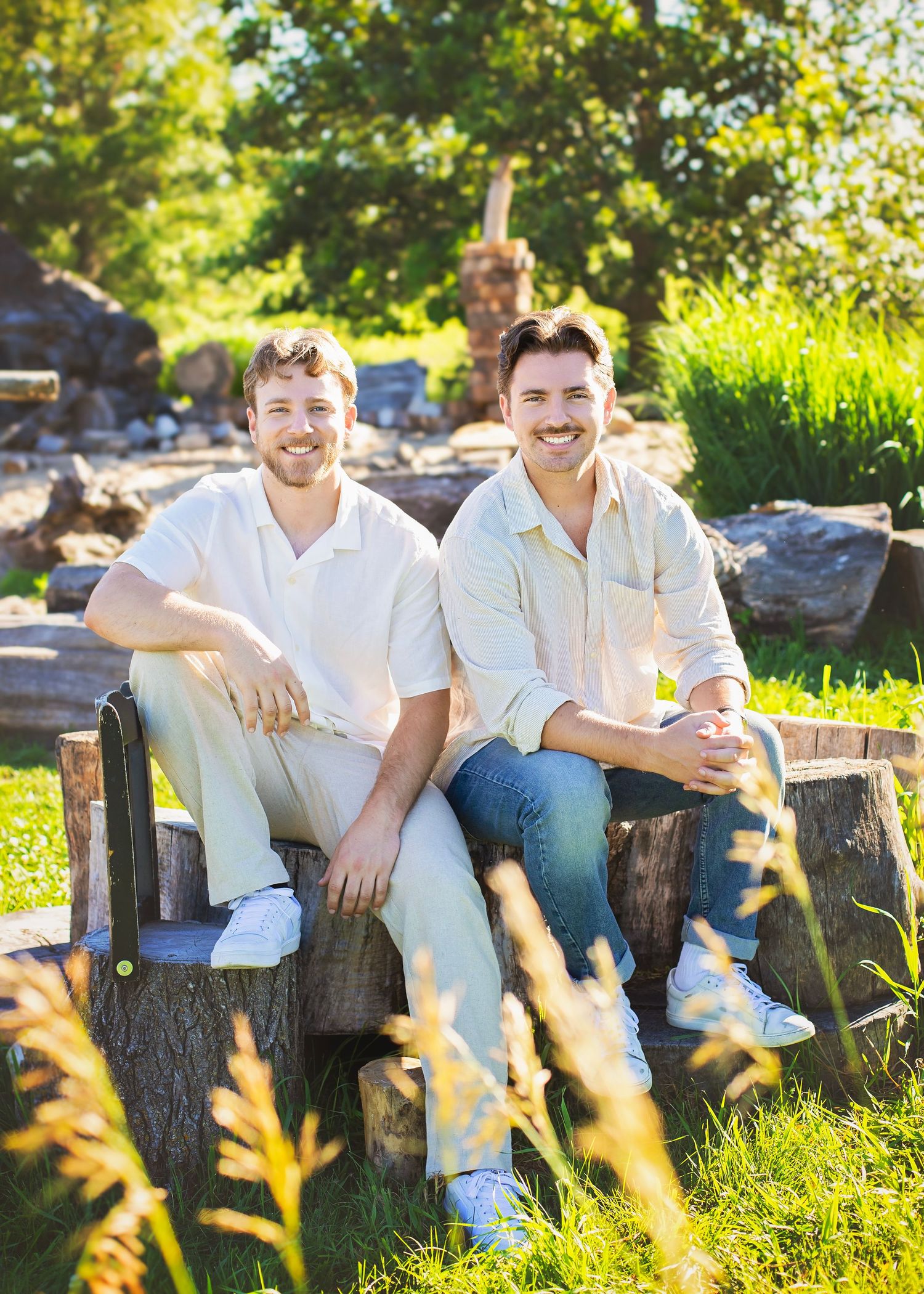Two smiling young men sit together on a rustic wooden bench in a sunny green garden setting.