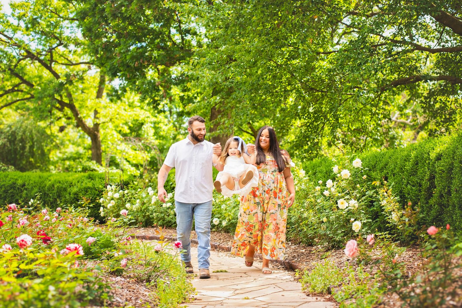 Family of three walks along a flower-lined garden path surrounded by lush greenery.
