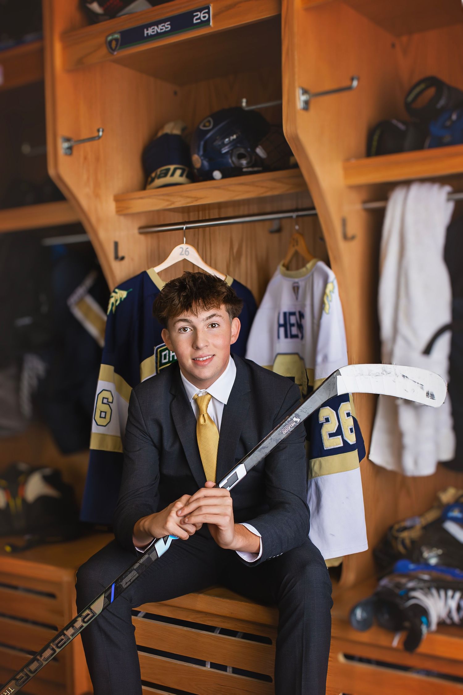 Young hockey player in suit holds stick in team locker room with jersey #26.