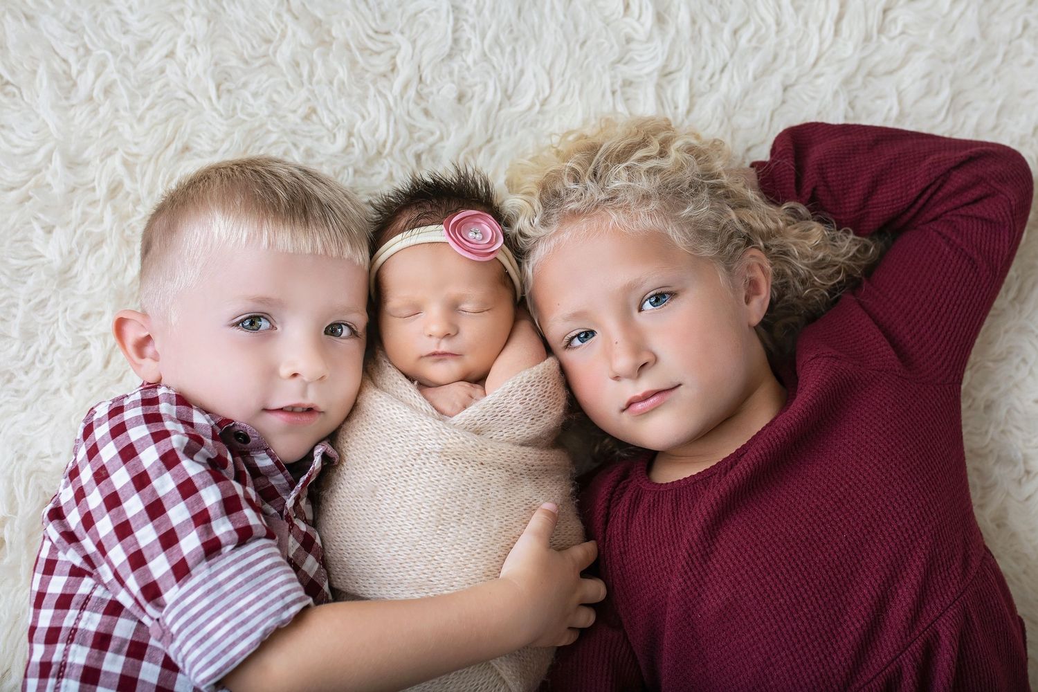 Two older siblings lying on white rug cuddling sleeping swaddled newborn baby with pink headband.