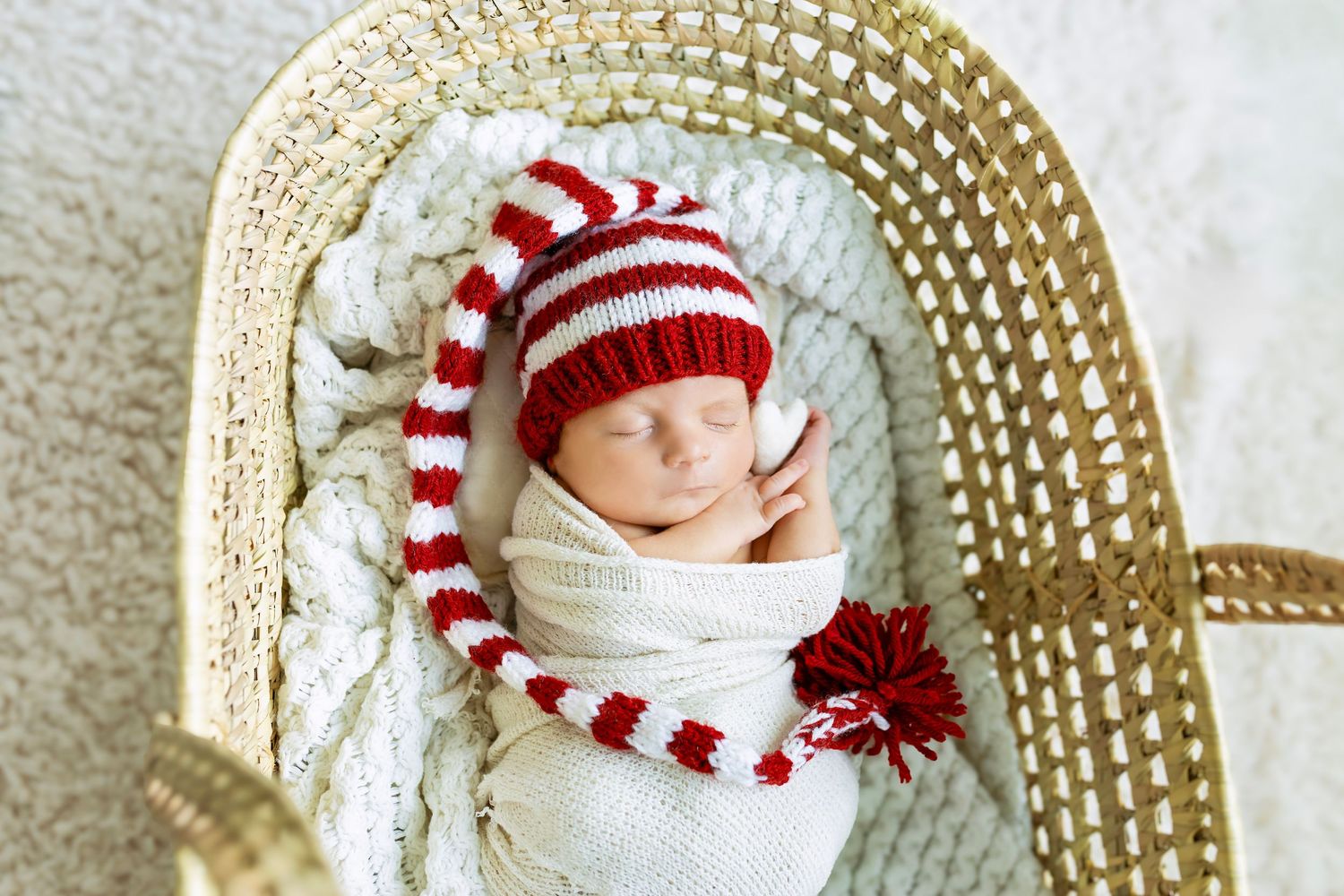 Sleeping newborn wrapped in white swaddle wearing red and white striped elf hat in wicker basket.