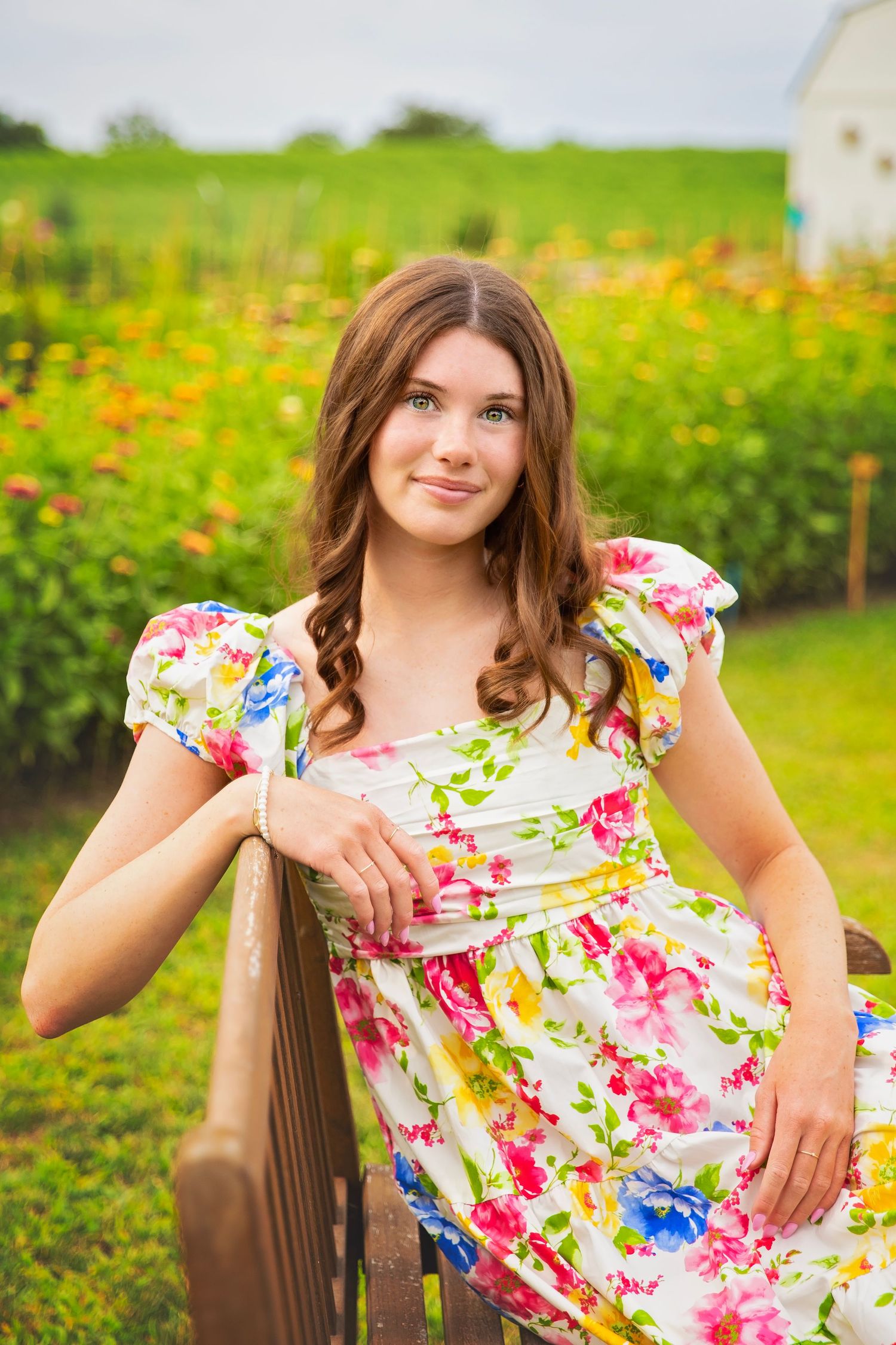 Teen girl in floral dress leaning on wooden fence in a colorful garden setting.