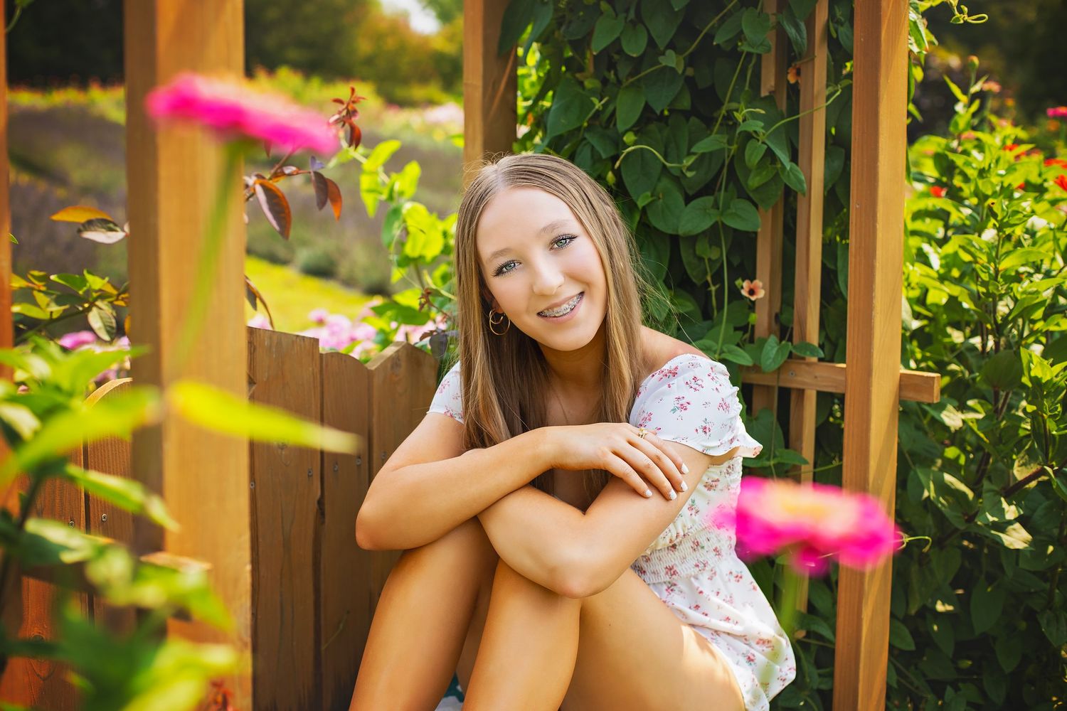 Smiling teenage girl in floral dress sitting with knees up in a sunny garden pergola.