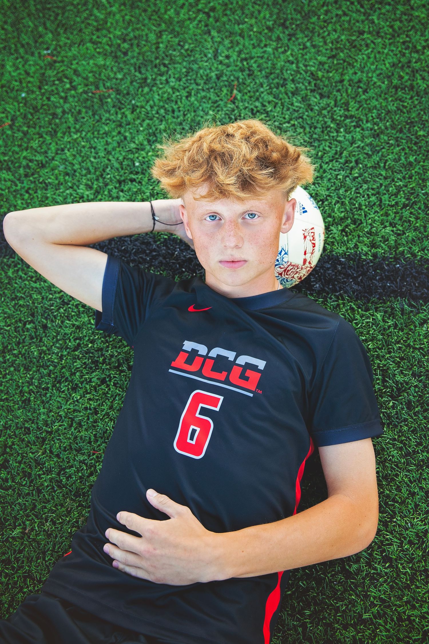 Teen soccer player wearing DCG number 6 jersey lying on green turf field with soccer ball.