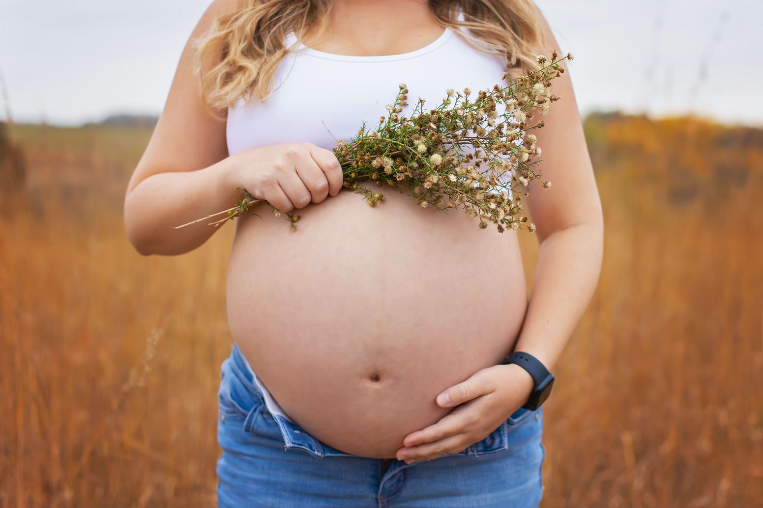 Close-up of pregnant woman's bare belly as she holds a small bouquet of wildflowers outdoors.