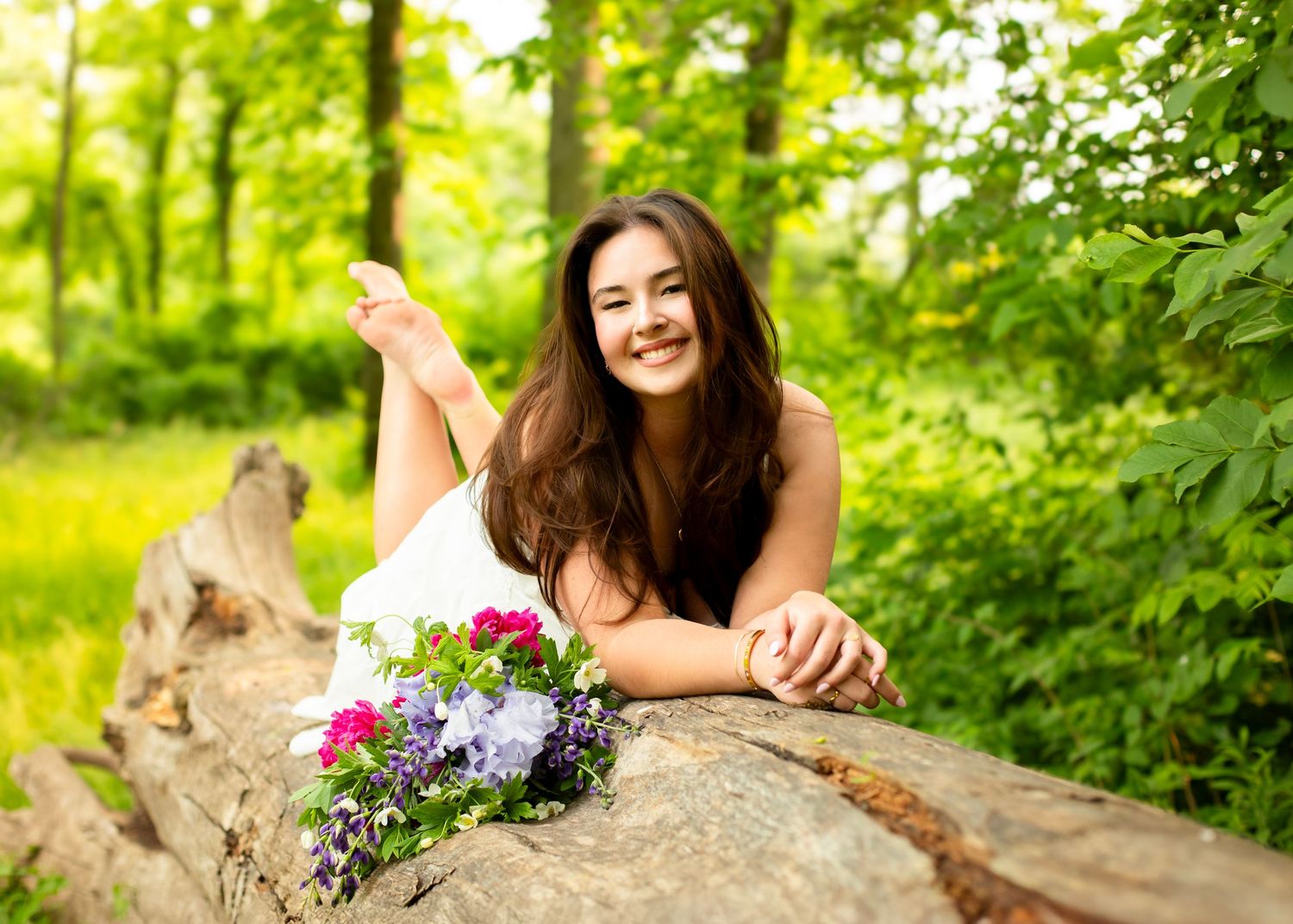 Smiling young woman in white dress rests on a log with a colorful bouquet in a sunny forest.
