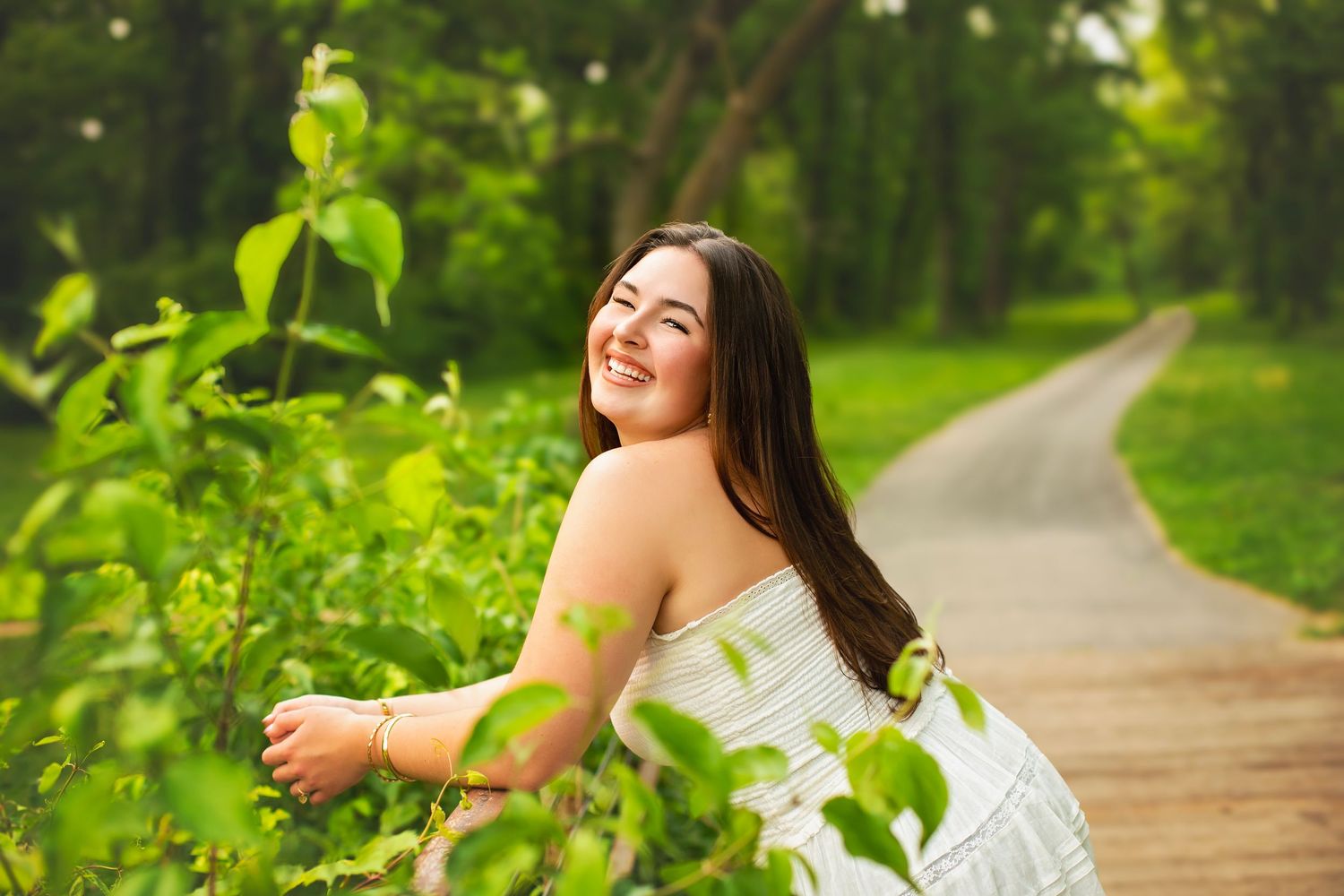 Smiling young woman in white dress leaning on green bushes along garden path.