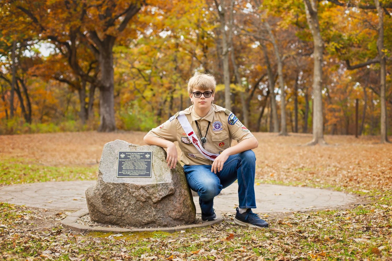 Teen in Boy Scout uniform crouching beside a memorial stone in an autumn park.